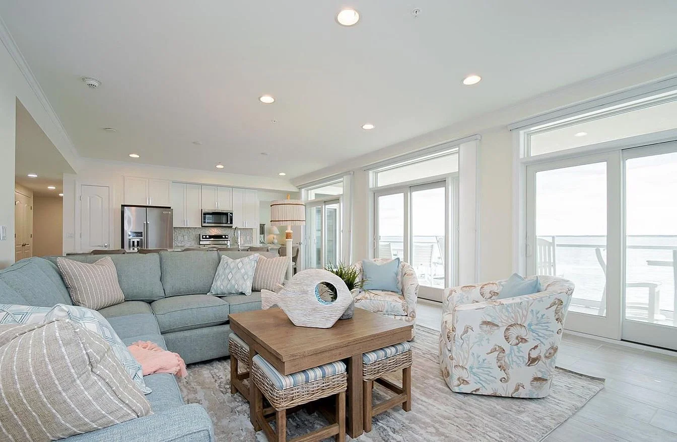Bright living room with light-colored furniture, coastal-themed armchairs, a wooden coffee table with decorative objects, and floor-to-ceiling windows with a view of a balcony and the ocean outside.
