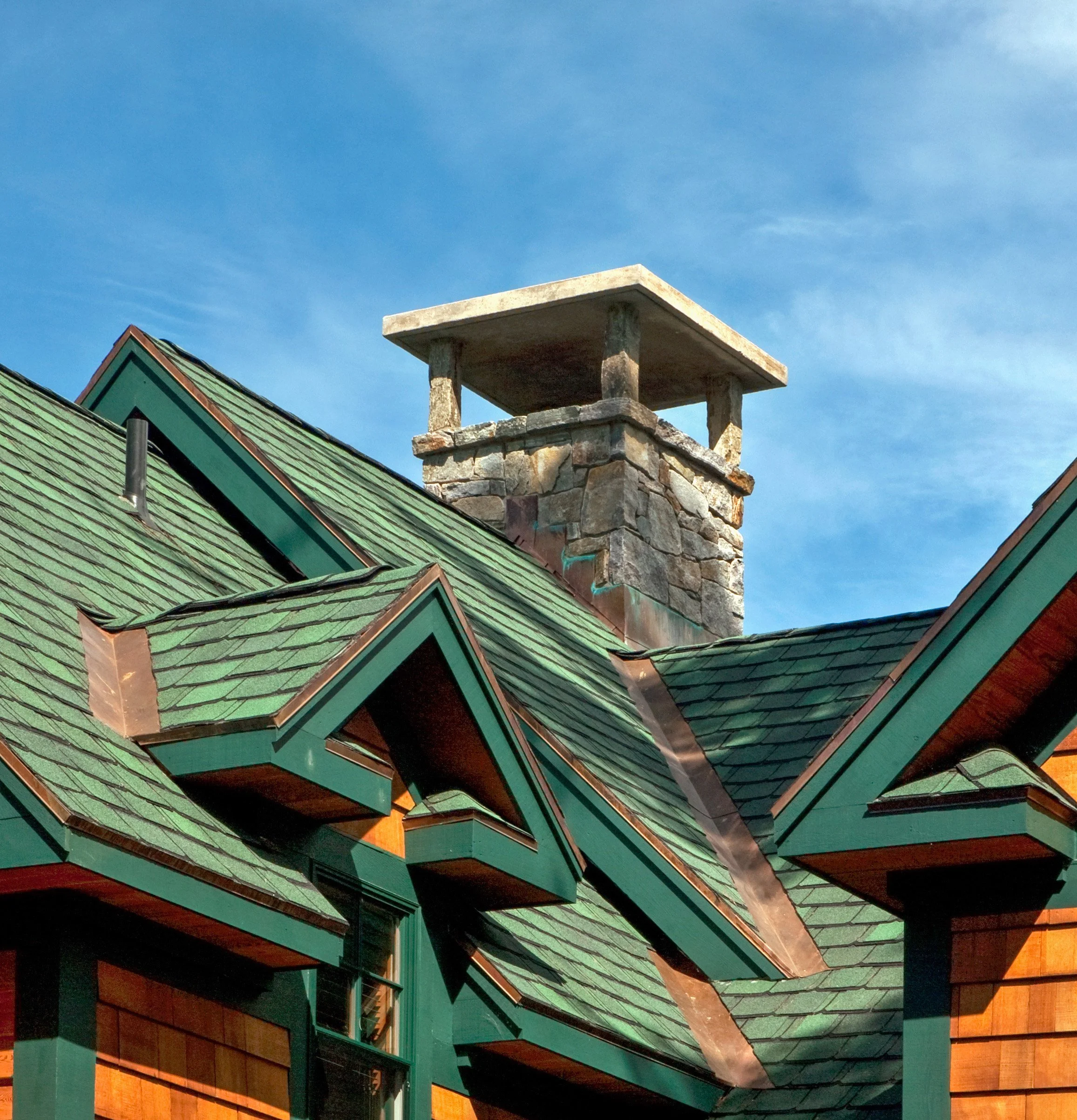 Close-up of a house roof with green shingles, dormer windows, and a stone chimney with a concrete cap, under a blue sky.