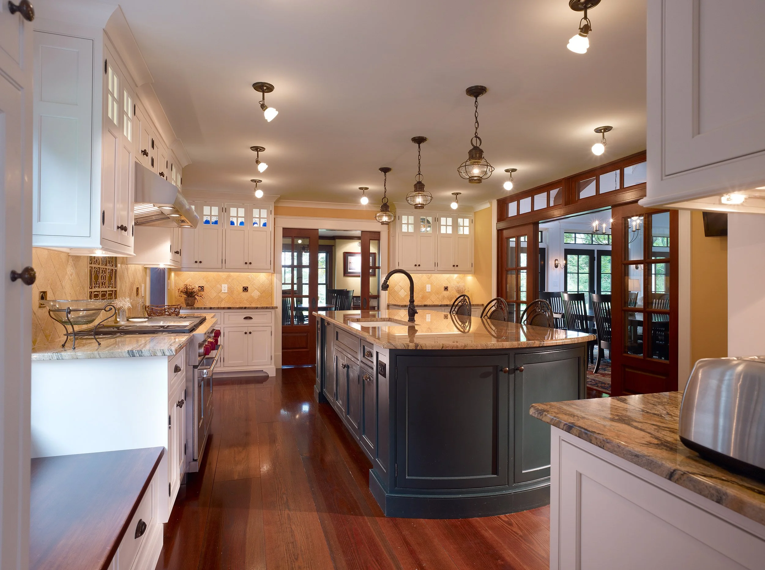 Bright kitchen with white cabinets, a large dark island with a granite countertop, pendant lights, and a view into an adjacent dining area with black chairs and large windows.