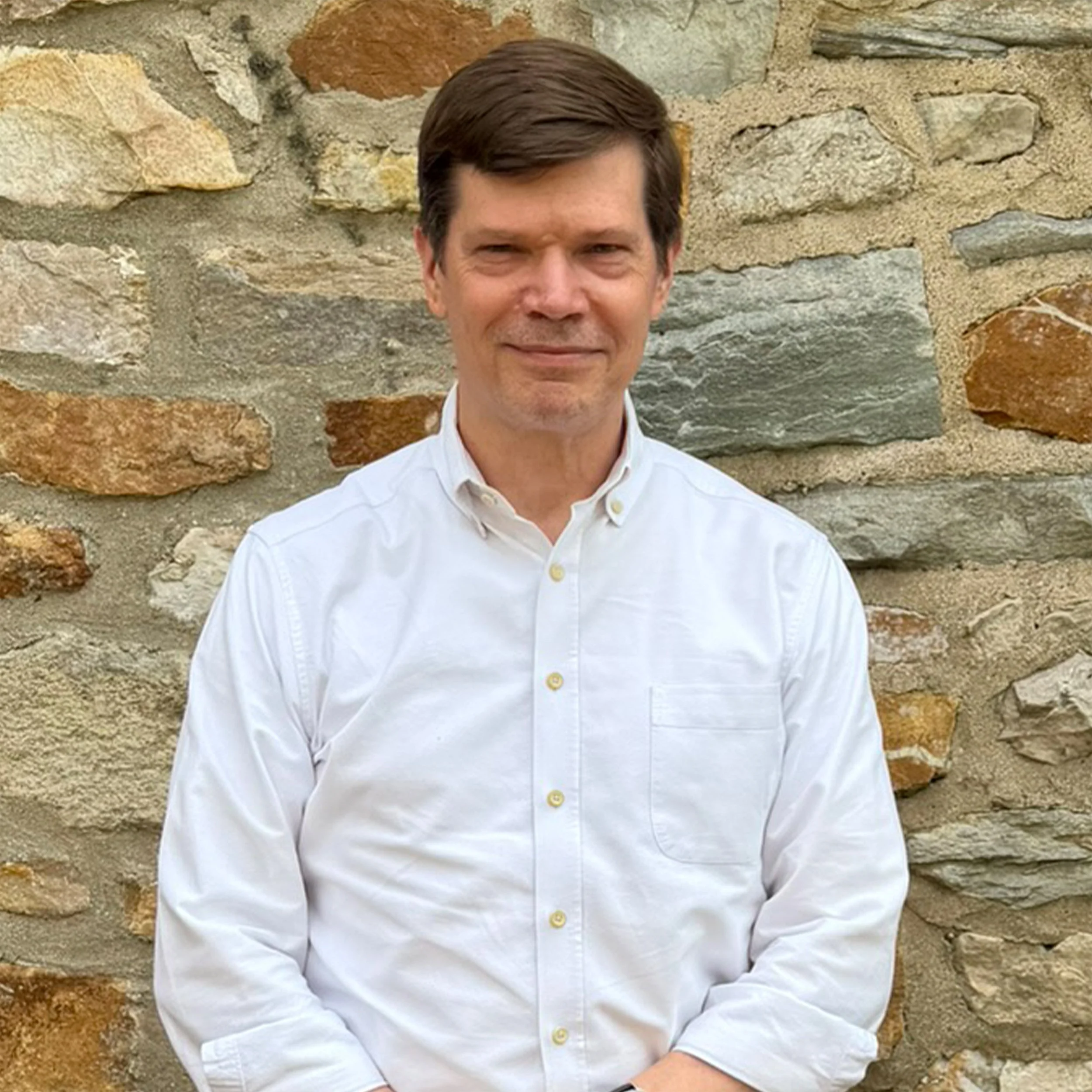 A man with brown hair wearing a white button-up shirt standing in front of a stone wall.