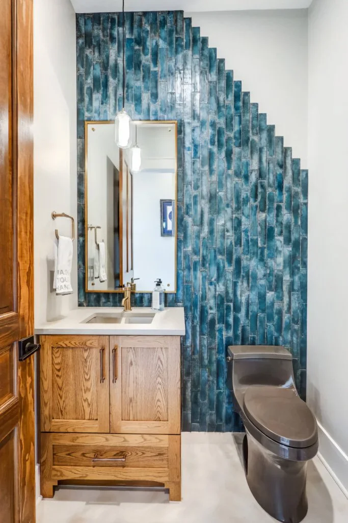 Modern bathroom with a wooden vanity, white countertop, and a large mirror. Blue textured tile wall behind the sink and toilet, with a white wall to the right. The toilet has a sleek, black design.