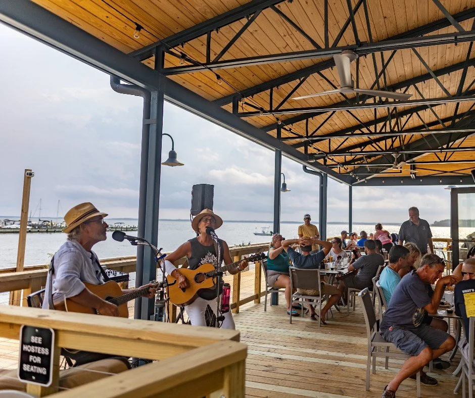 Musicians playing guitars on a covered outdoor patio overlooking a body of water with people dining and boats in the background.