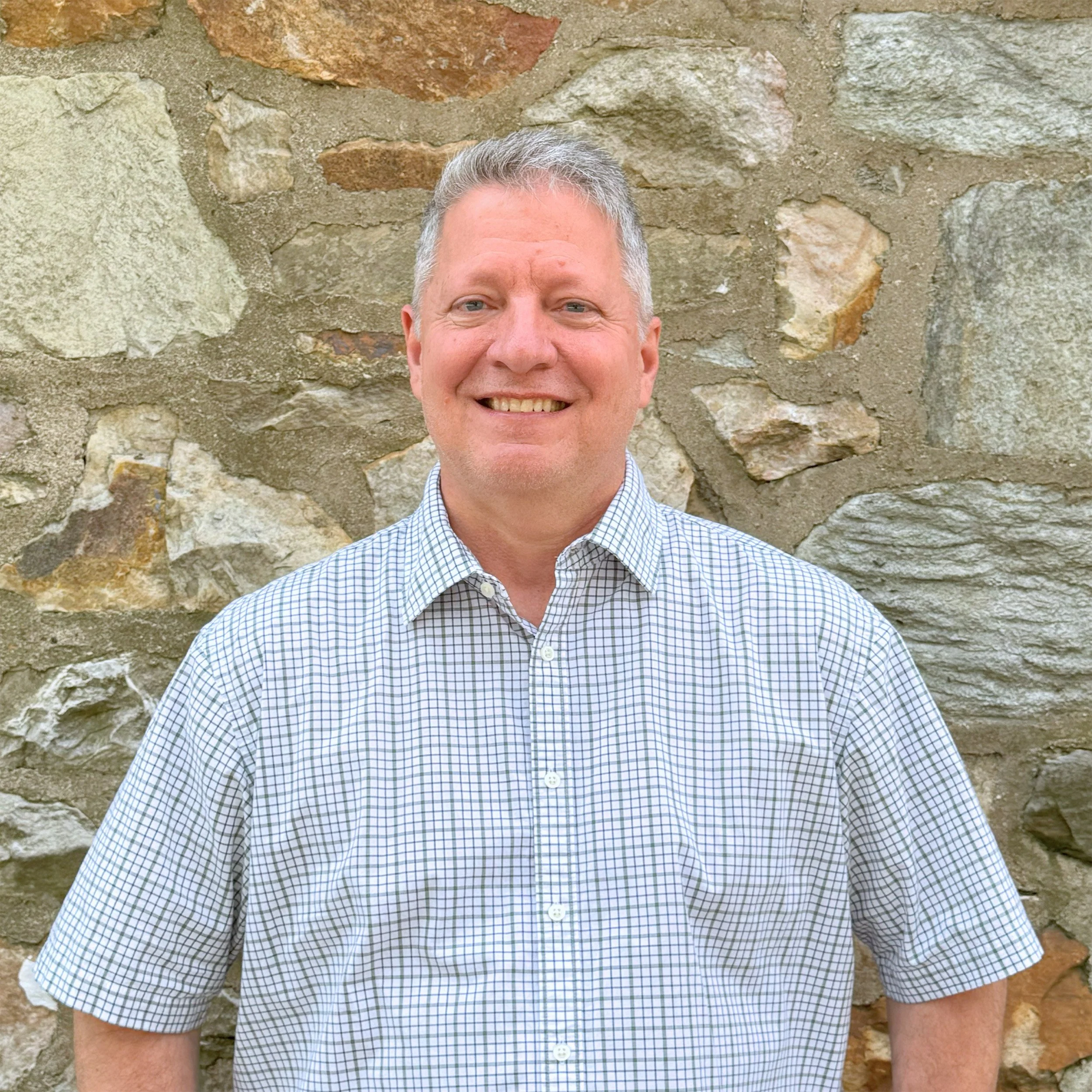 A smiling man in a blue and white checkered shirt standing in front of a stone wall.