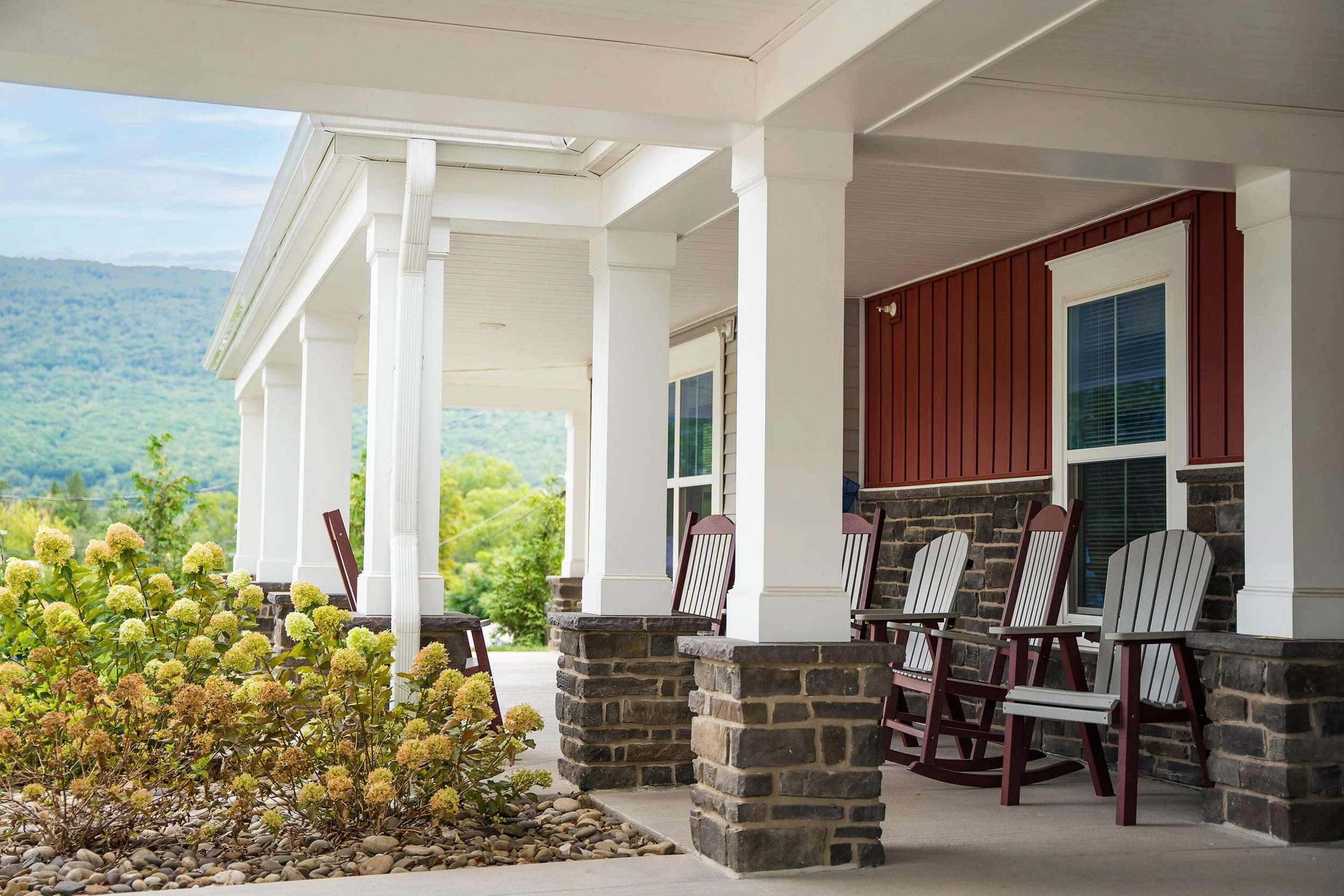 Porch of a residential building with multiple rocking chairs, white pillars, stone base, flower bed, and a view of green hills in the background.