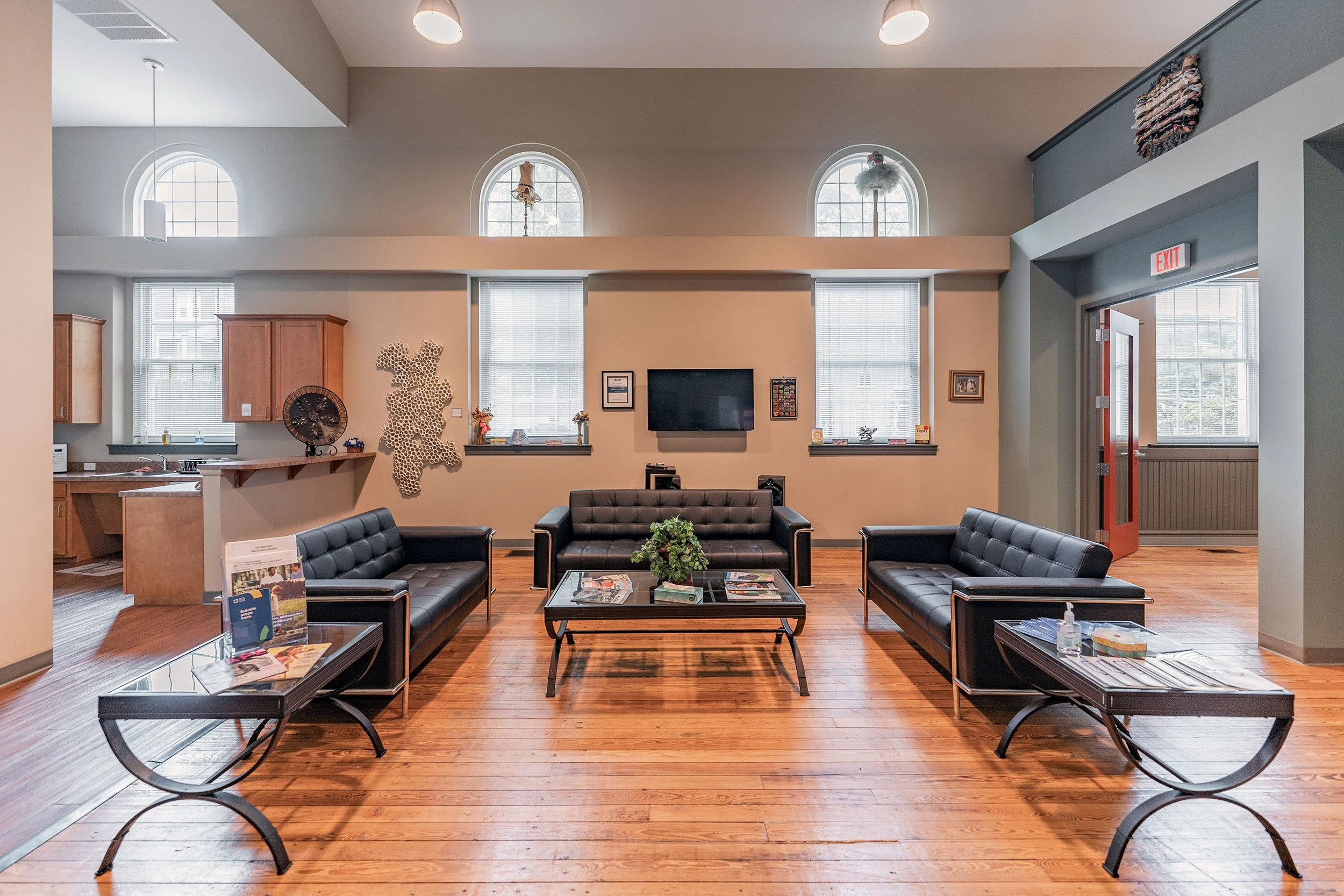 A spacious lounge with three black sofas, a coffee table with a plant, wooden floors, and a wall-mounted TV with three windows above, decorated with art and plants. The kitchen area is visible on the left, and an exit door on the right.