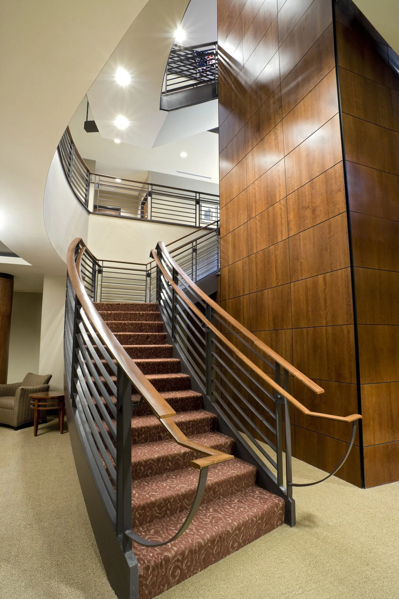 Interior view of a staircase with wooden handrails, brown patterned carpet, and a curved wooden wall in a building with multiple floors and ceiling lights.
