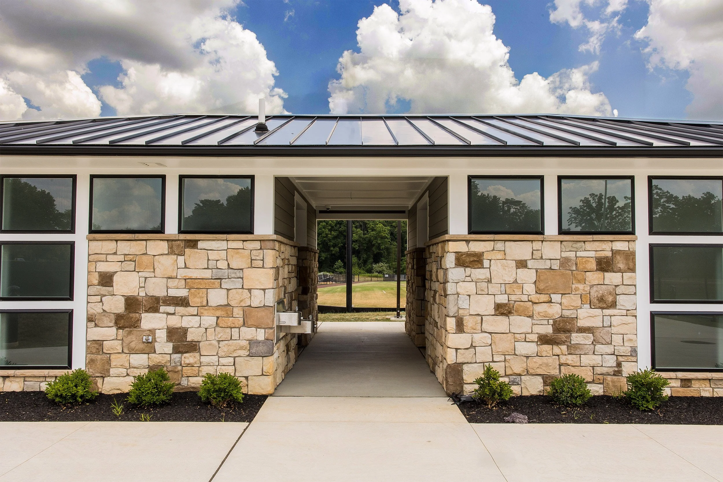 Front view of a restroom building with stone walls, black window frames, and a metal roof, featuring an open walkway in the center leading to a grassy field with trees in the background, under a partly cloudy sky.