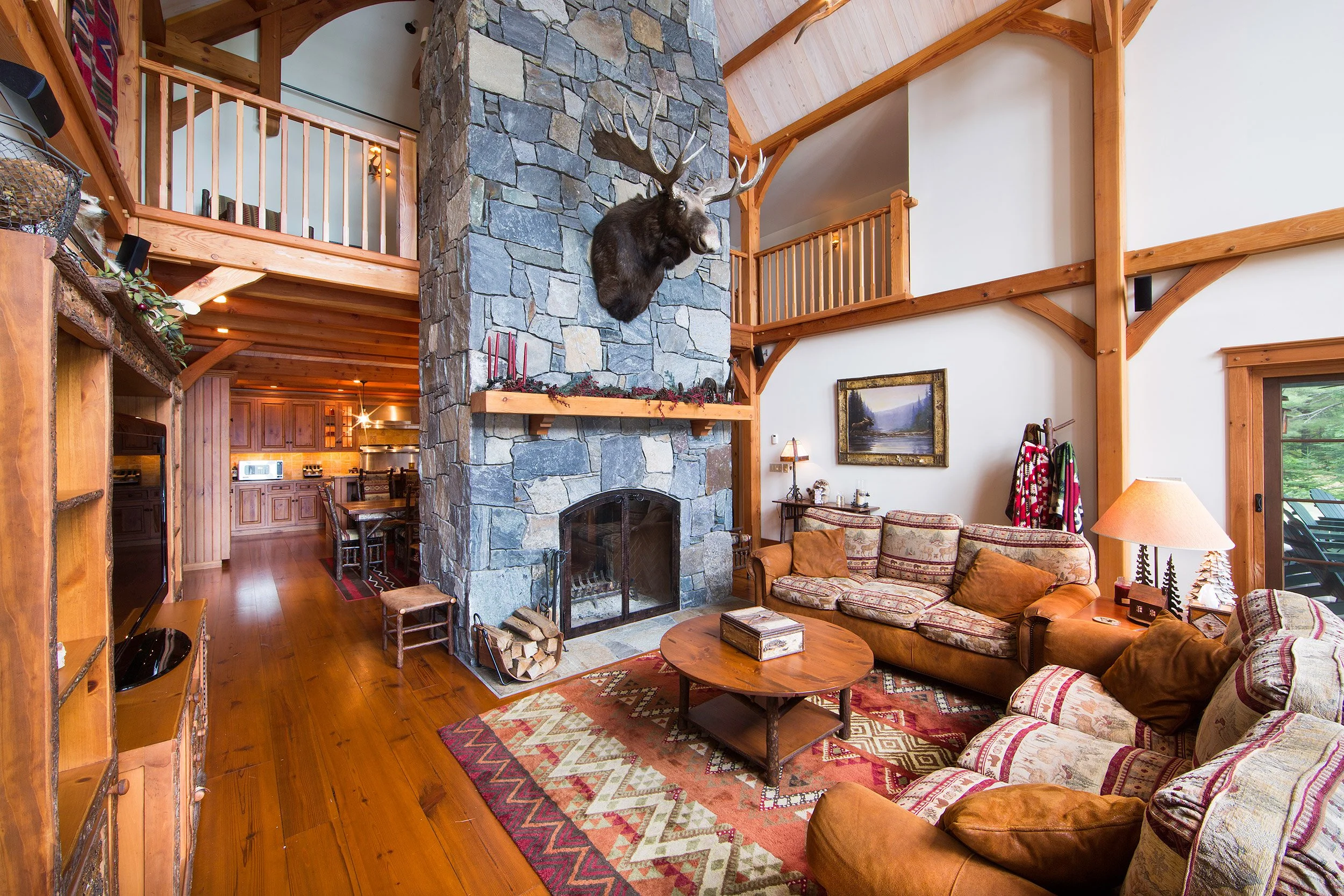 Cozy living room in an Adirondack style home with a stone fireplace, a mounted moose head, wooden beams and railings, a patterned area rug, floral and leather sofas, a wooden coffee table, lamps, and a large window.