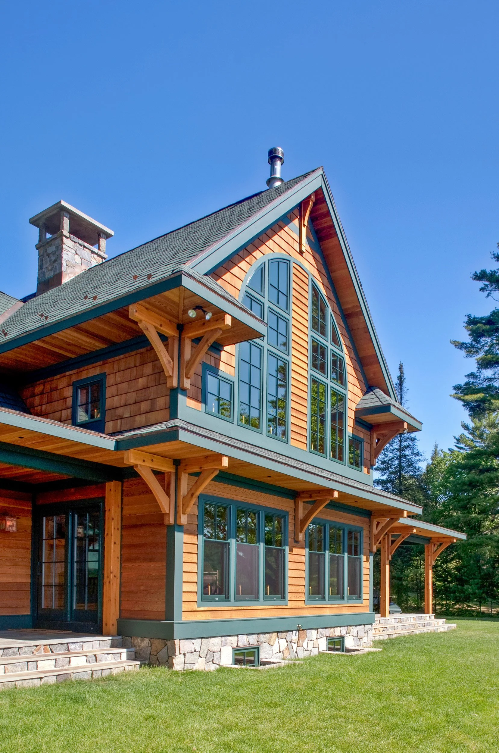 A two-story Adirondack style house with wooden siding, large windows, and a stone foundation, surrounded by a green lawn and trees, under a clear blue sky.
