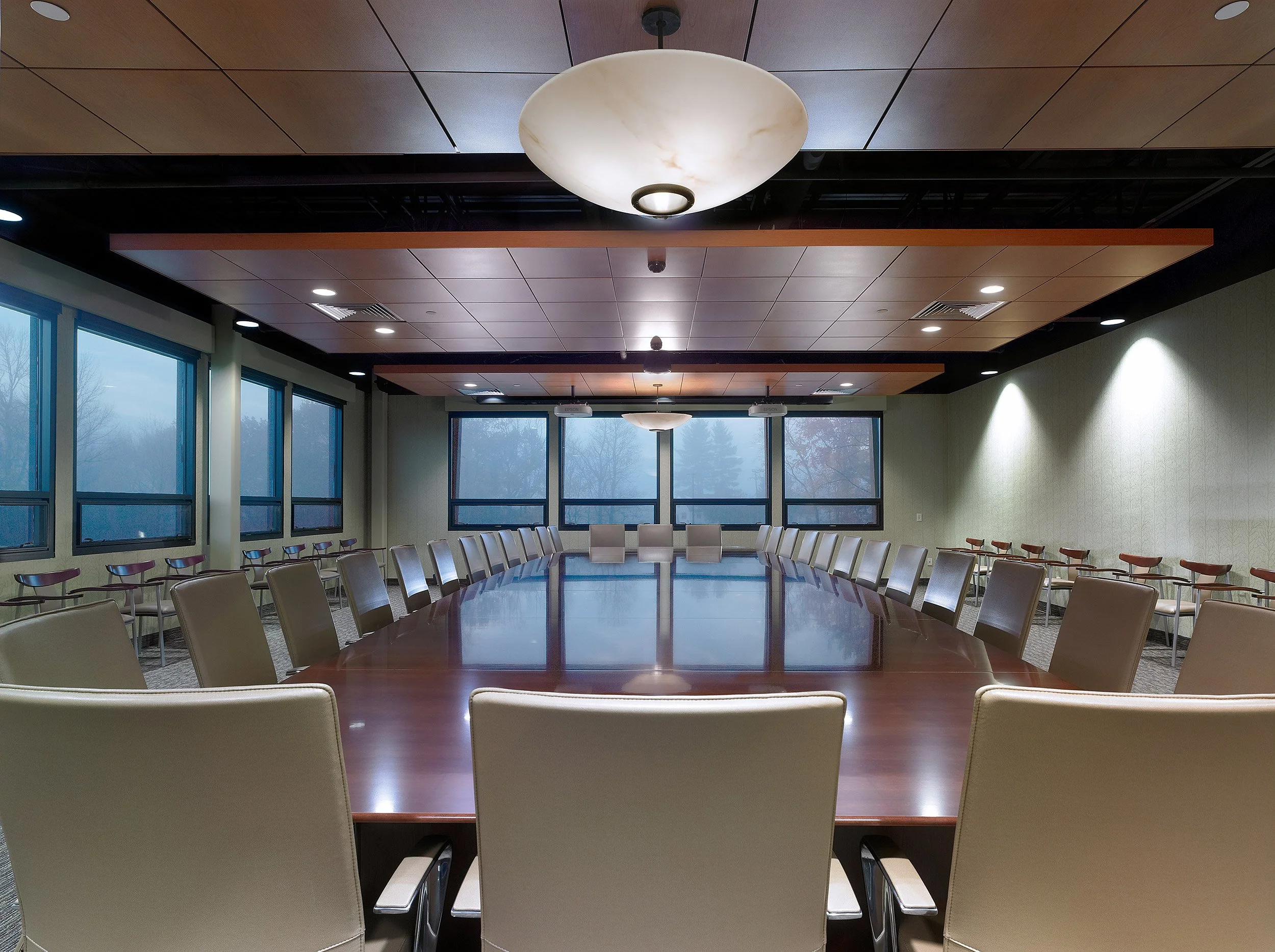 Empty conference room with a large wooden table, beige chairs, and overhead lighting. Windows on one side showing a cloudy outdoor scene.