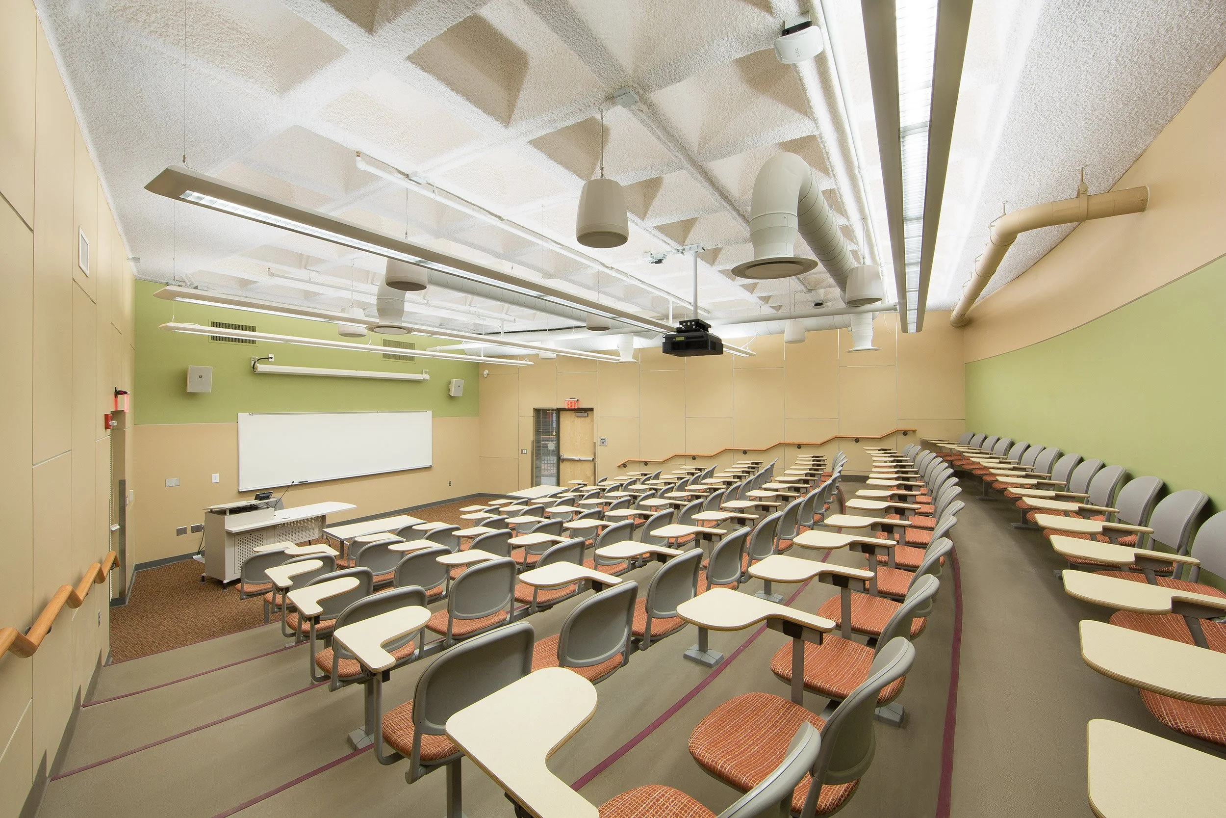 Empty college classroom with tiered seating, a whiteboard, and a projector mounted on the ceiling.