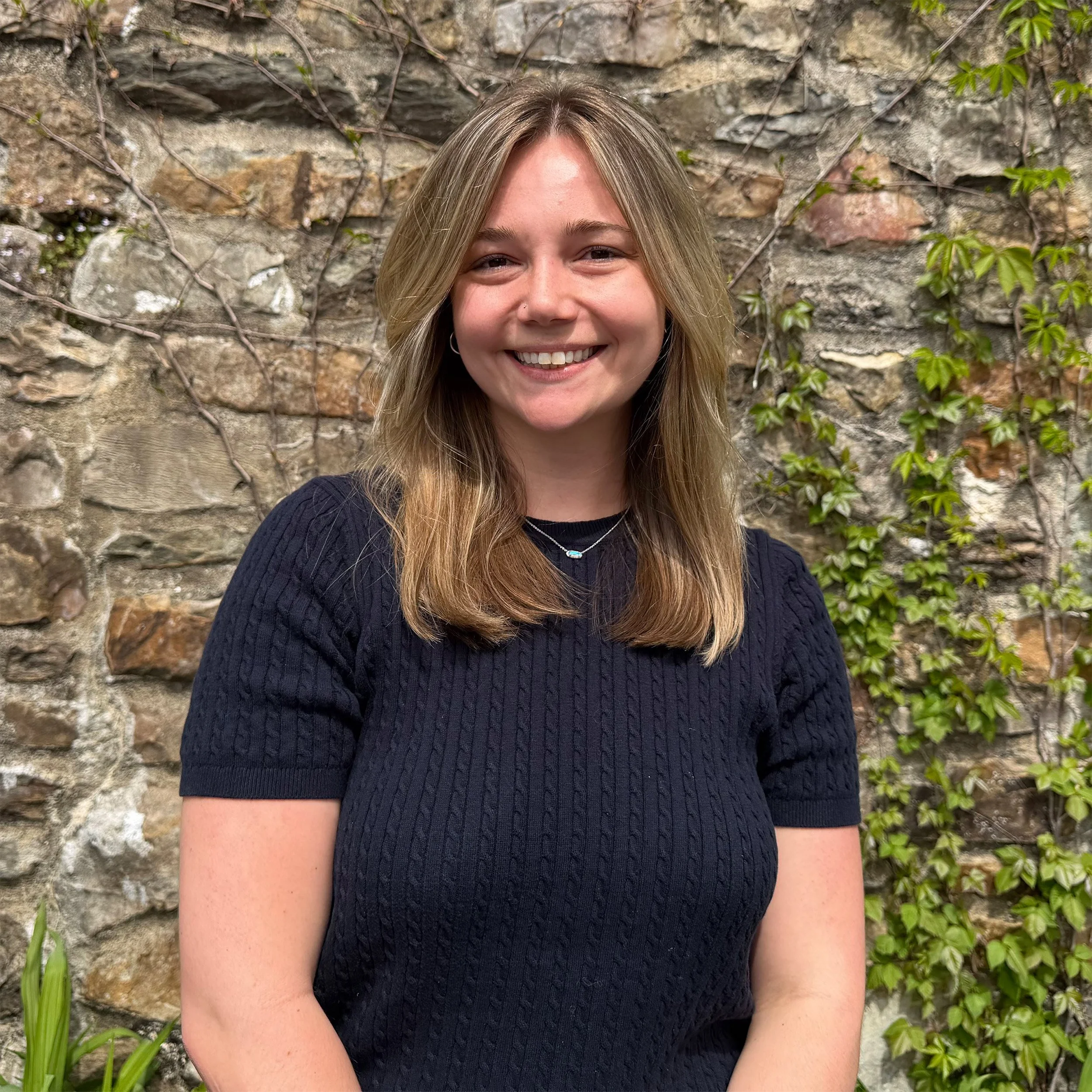 A woman with blond hair smiling in front of a stone wall with green ivy and plants.