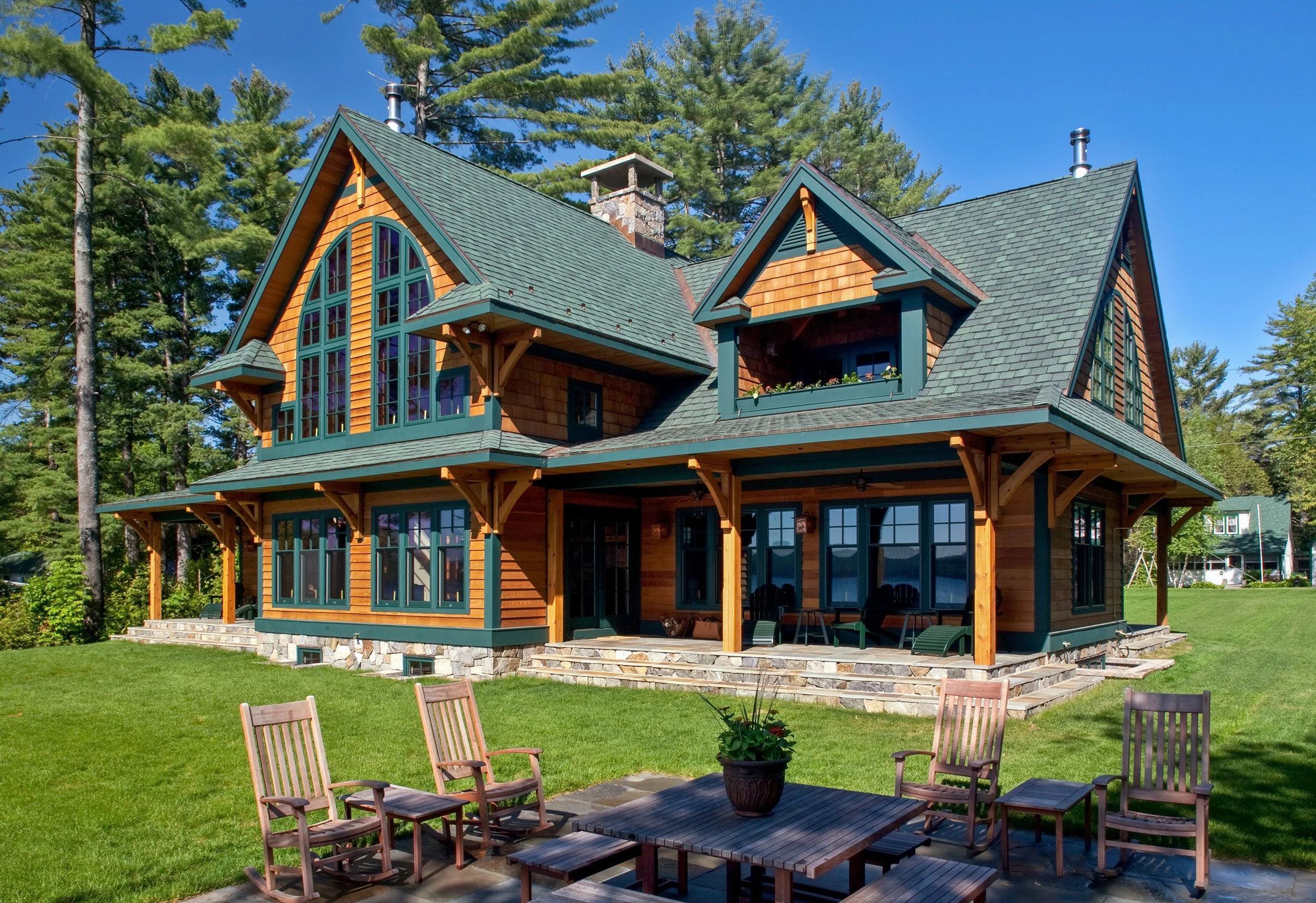 A large two-story Adirondack style lakehouse with wooden siding and multiple green gabled roofs, surrounded by tall trees, with a spacious grassy lawn and a patio with outdoor seating and a potted plant.