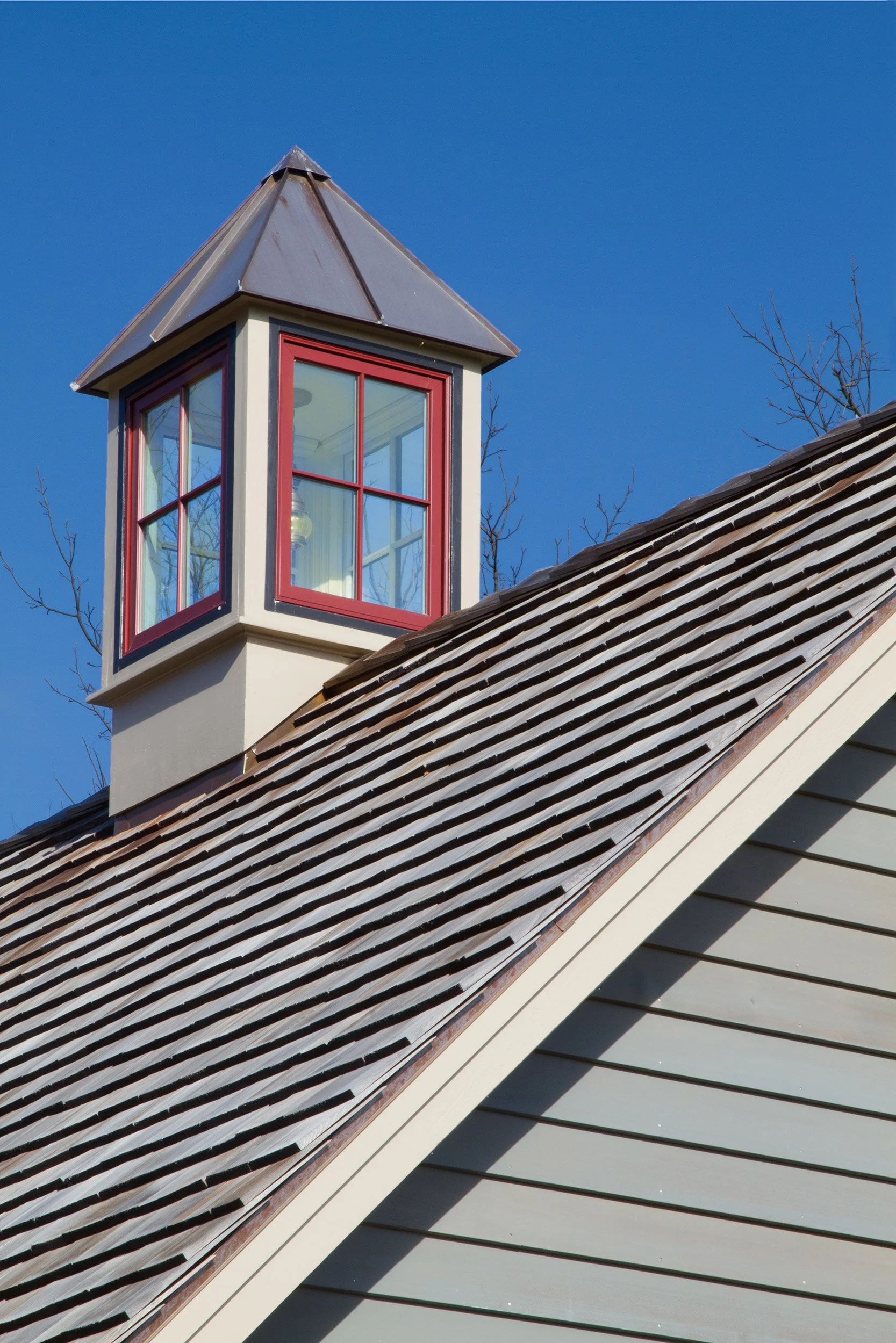 Close-up of a house roof with brown shingles and a side windowed cupola with red trim against a clear blue sky.
