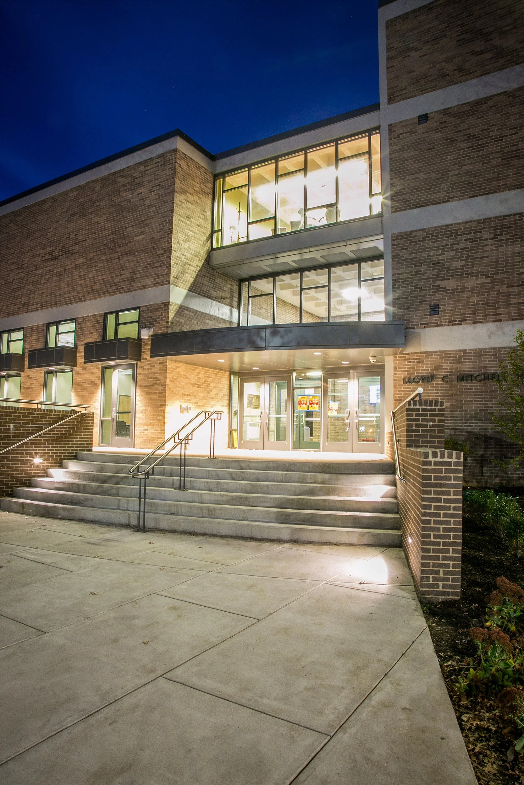 Nighttime photo of the exterior of Lloyd G. Mitchell academic building with brick walls, glass doors, and illuminated windows, with a set of stairs and railings leading to the entrance.