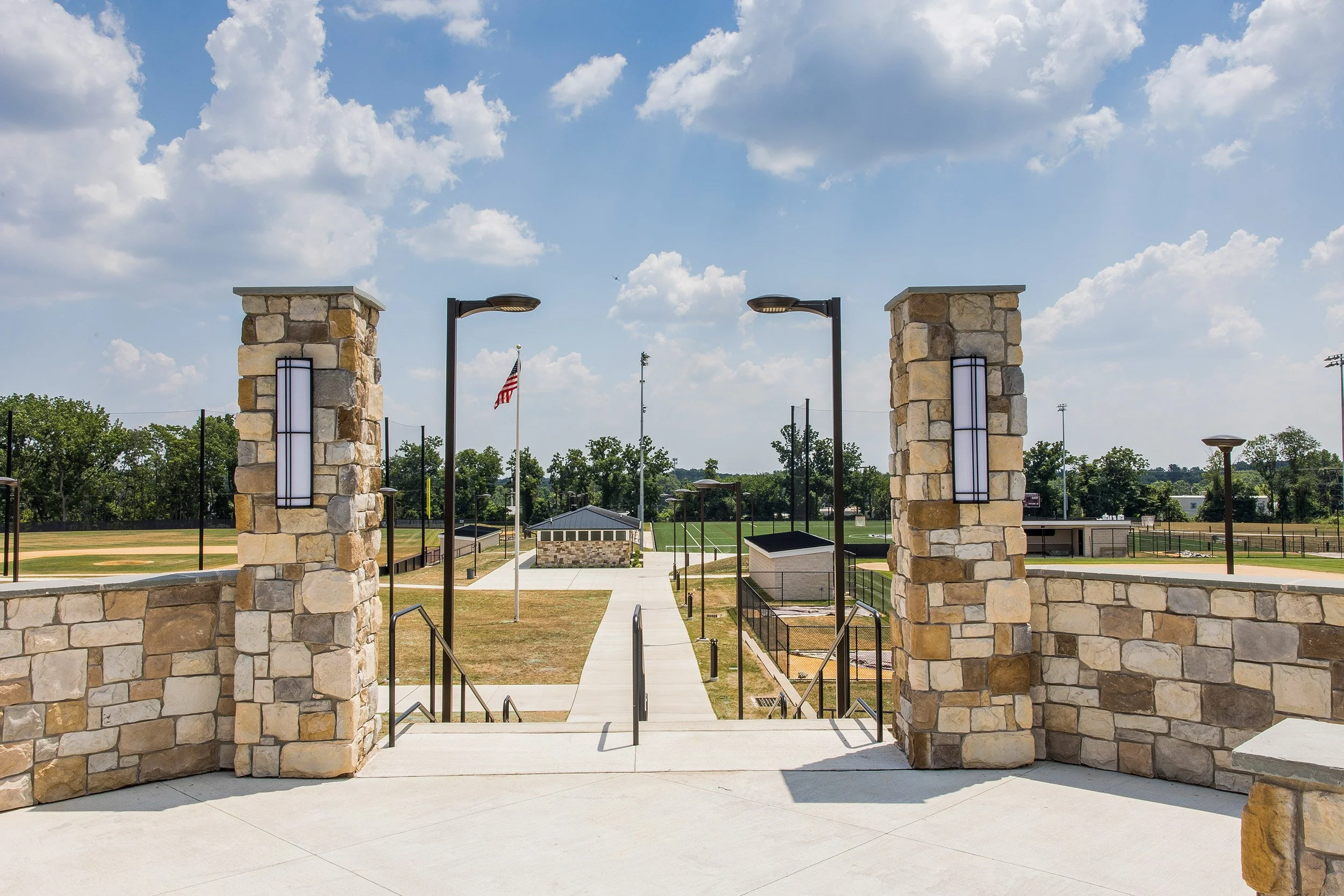Entrance to an athletic complex with stone pillars, a concrete walkway, and lamp posts, with sports fields and trees in the background under a partly cloudy sky.