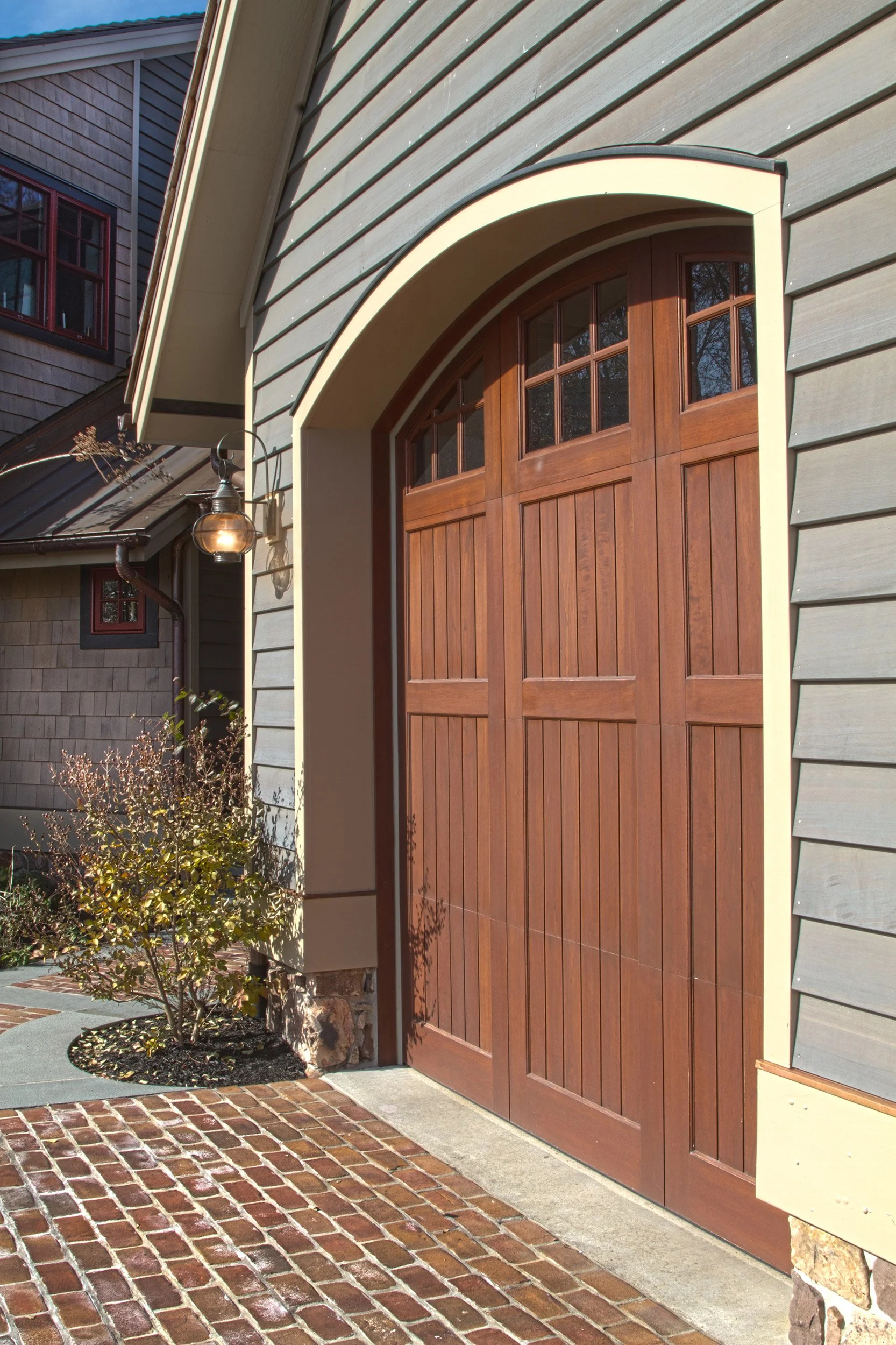 Wooden garage door with small windows at the top, attached to a house with grey siding and a landscaped brick driveway.