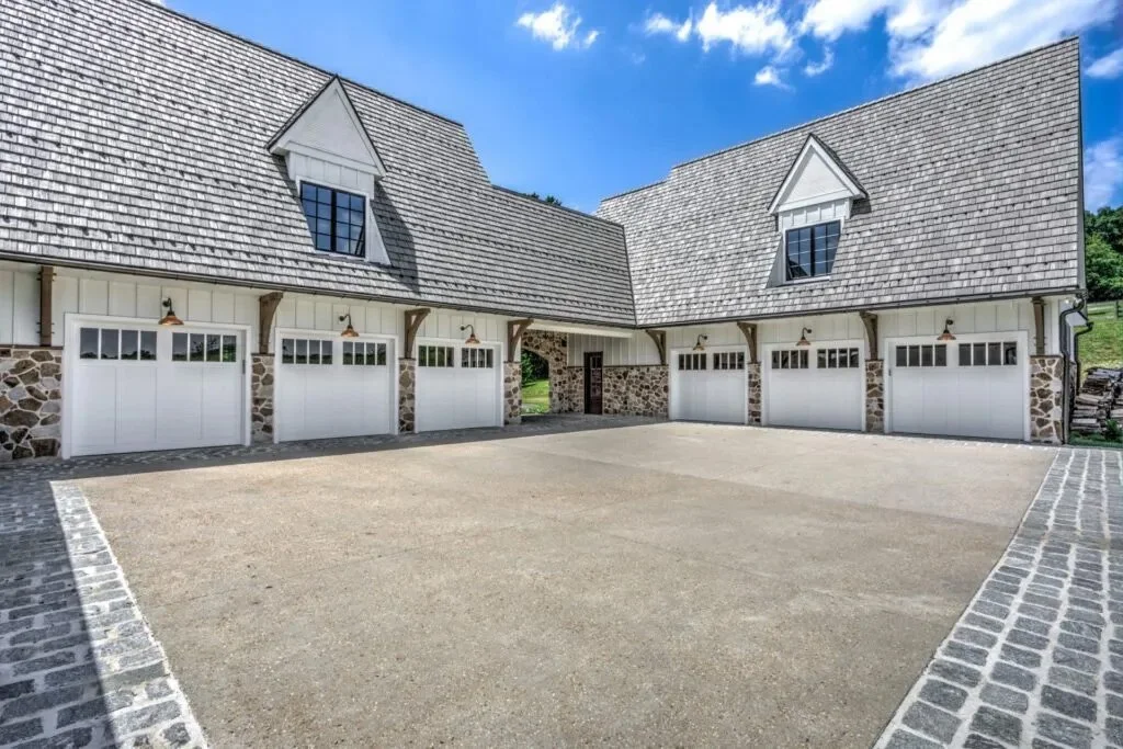 A large courtyard with a gravel driveway, surrounded by a stone and white wooden garage building, under a partly cloudy blue sky.
