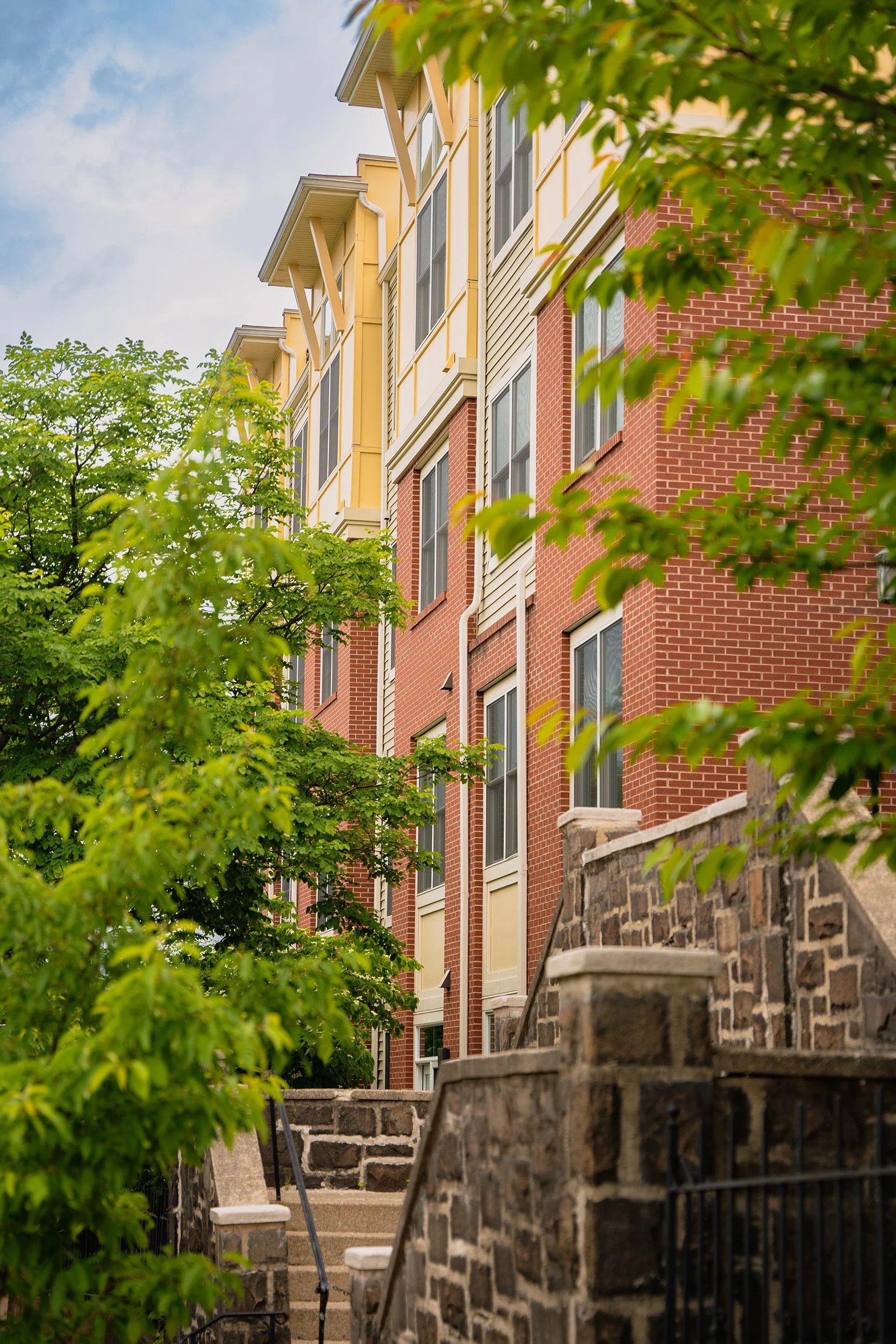 View of an apartment building with brick and siding exterior, surrounded by green trees, with stone stairs leading up to the entrance.