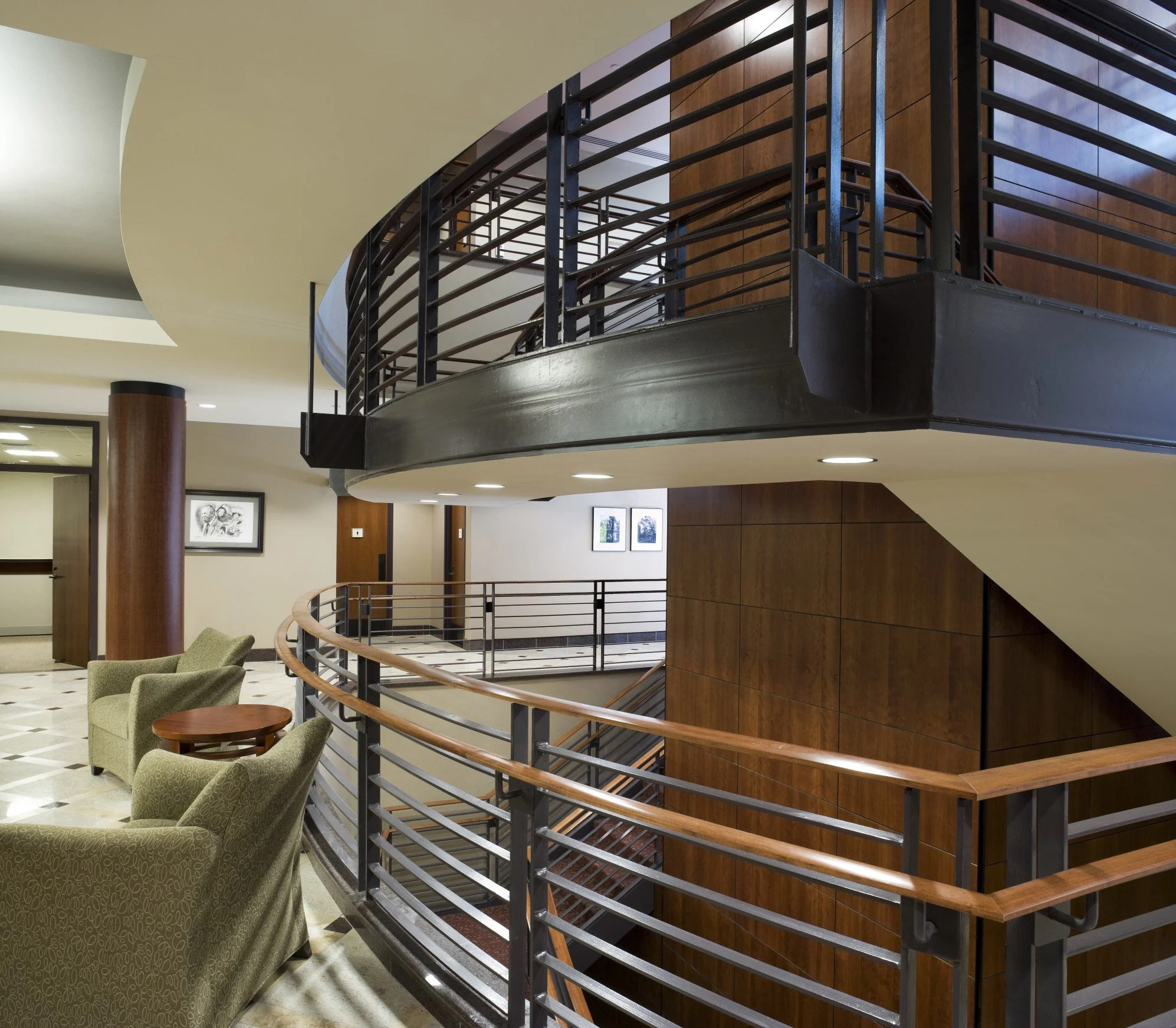 Interior of a modern building with a curved staircase, wooden handrails, metal railings, and seating area with armchairs and small table.