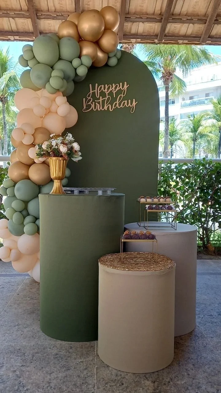 Décoration d'anniversaire avec un panneau vert, ballons de différentes tailles en couleurs or, vert et beige, une urne à fleurs, et des plateaux avec des cupcakes, en intérieur avec vue sur un jardin tropical.