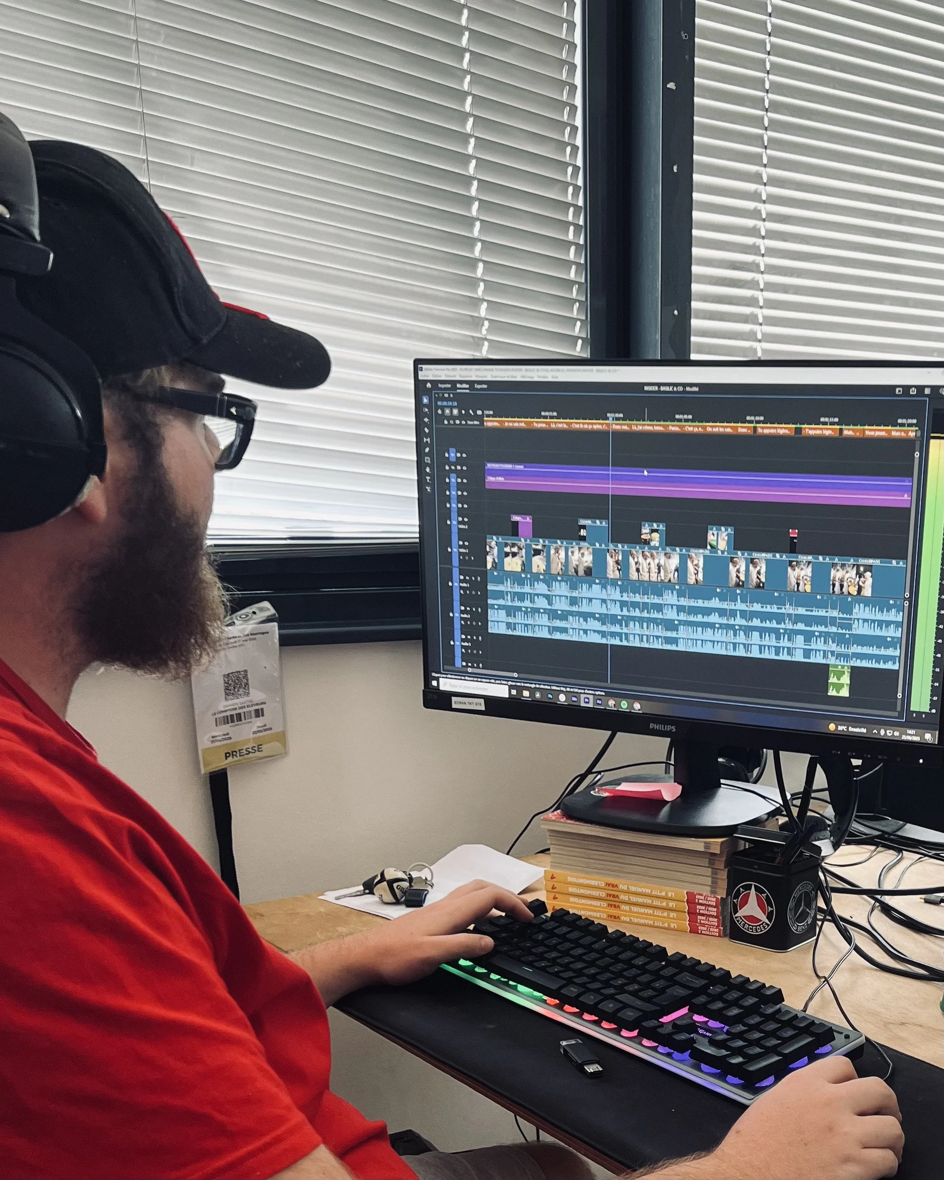 Un homme avec une barbe, portant un casque audio et une casquette noire, travaille sur un ordinateur avec un écran affichant un logiciel de montage vidéo, dans une pièce avec des stores enrouleurs et un badge presse accroché à la fenêtre.