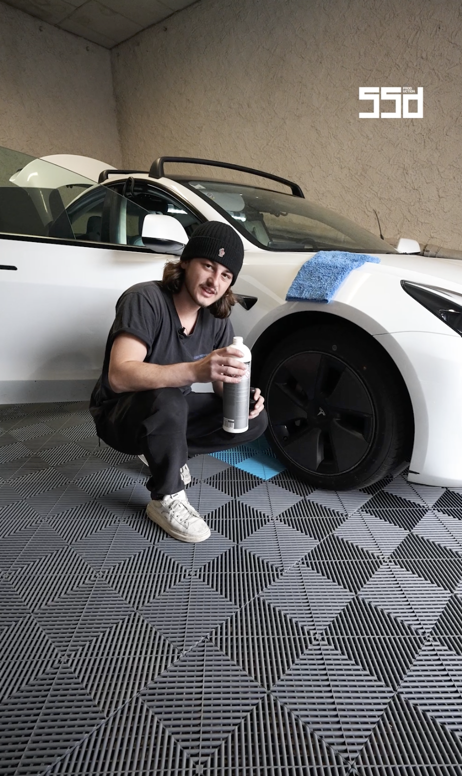 Un homme en sweat noir, avec un bonnet noir, tenant une bouteille de produit pour lavage de voiture, plié devant une Tesla blanche dans un garage, en train de nettoyer la roue avec un tapis de sol en grilles.