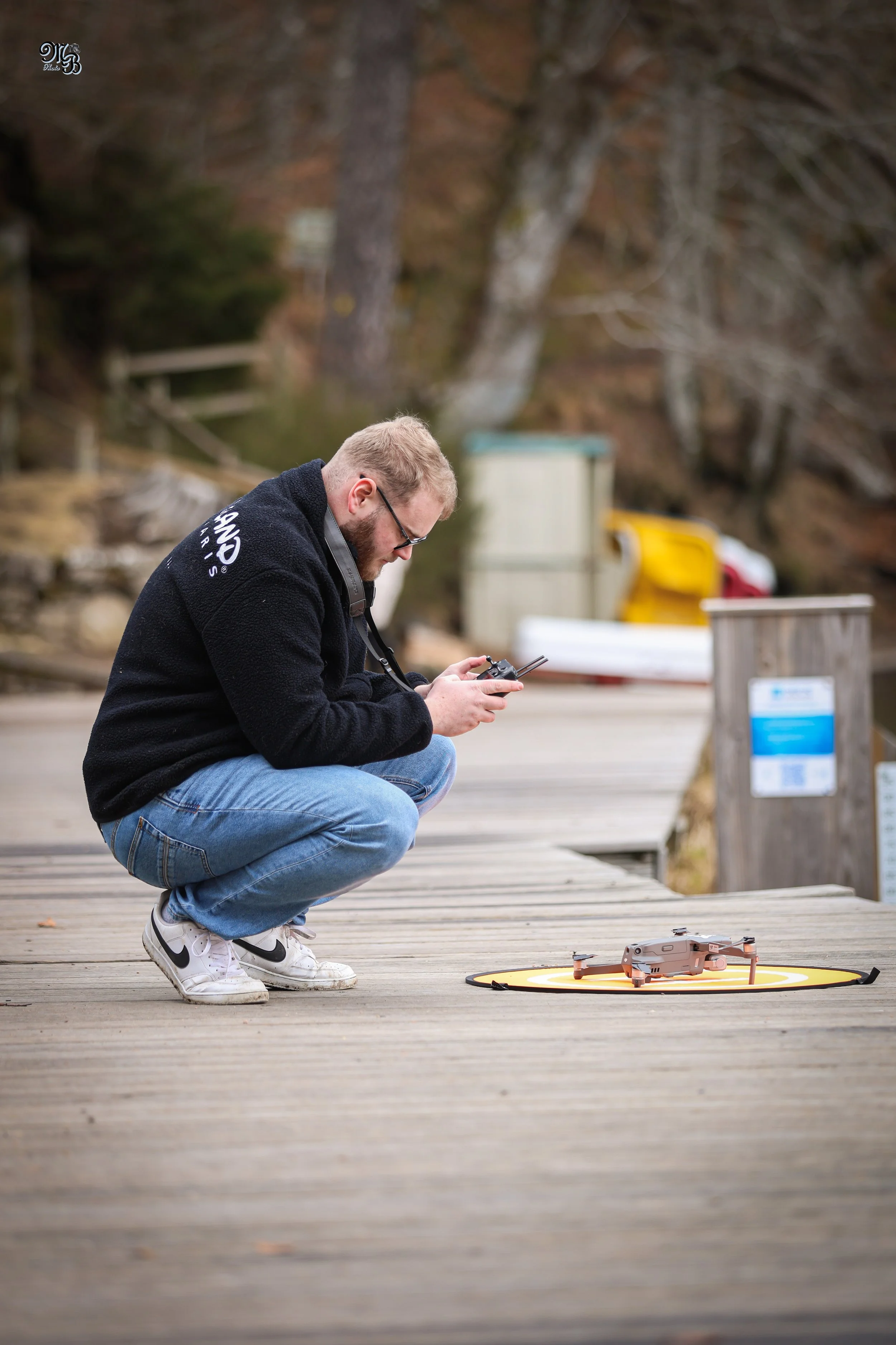 Un homme en position accroupie, portant des vêtements décontractés, regarde un drone posé au sol sur un mat indiqué par un cercle orange. Il semble préparer le drone pour un vol, dans un environnement extérieur avec des arbres et des cailloux en arrière-plan.