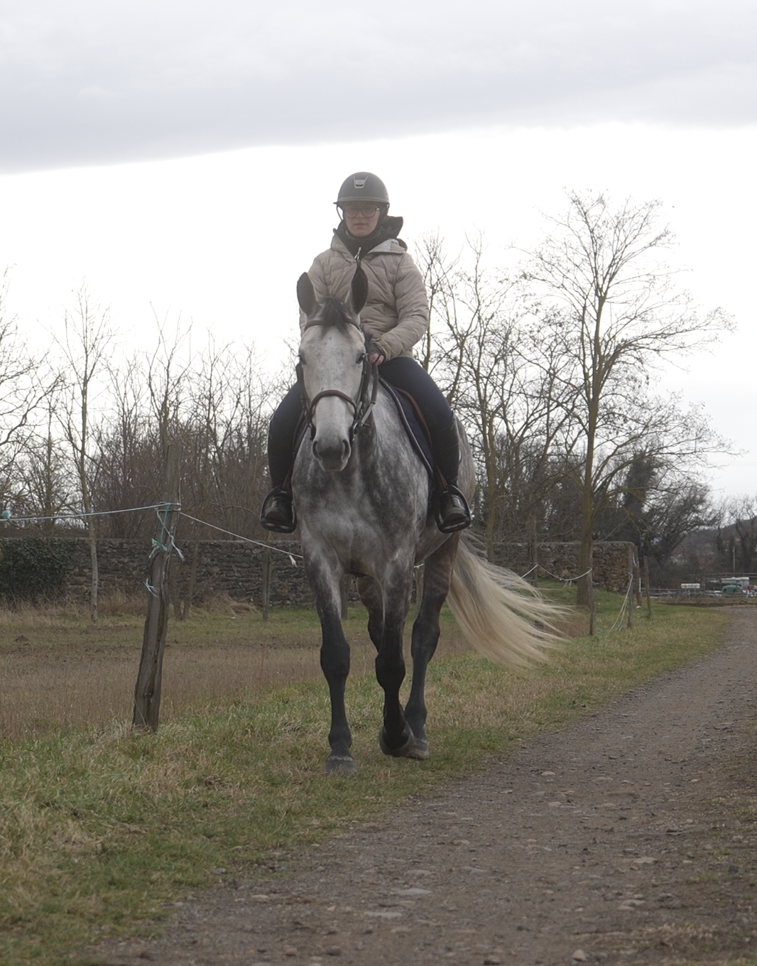 Jeune femme à cheval dans un chemin rural, portant un casque, veste beige et lunettes, avec des arbres sans feuilles en arrière-plan.