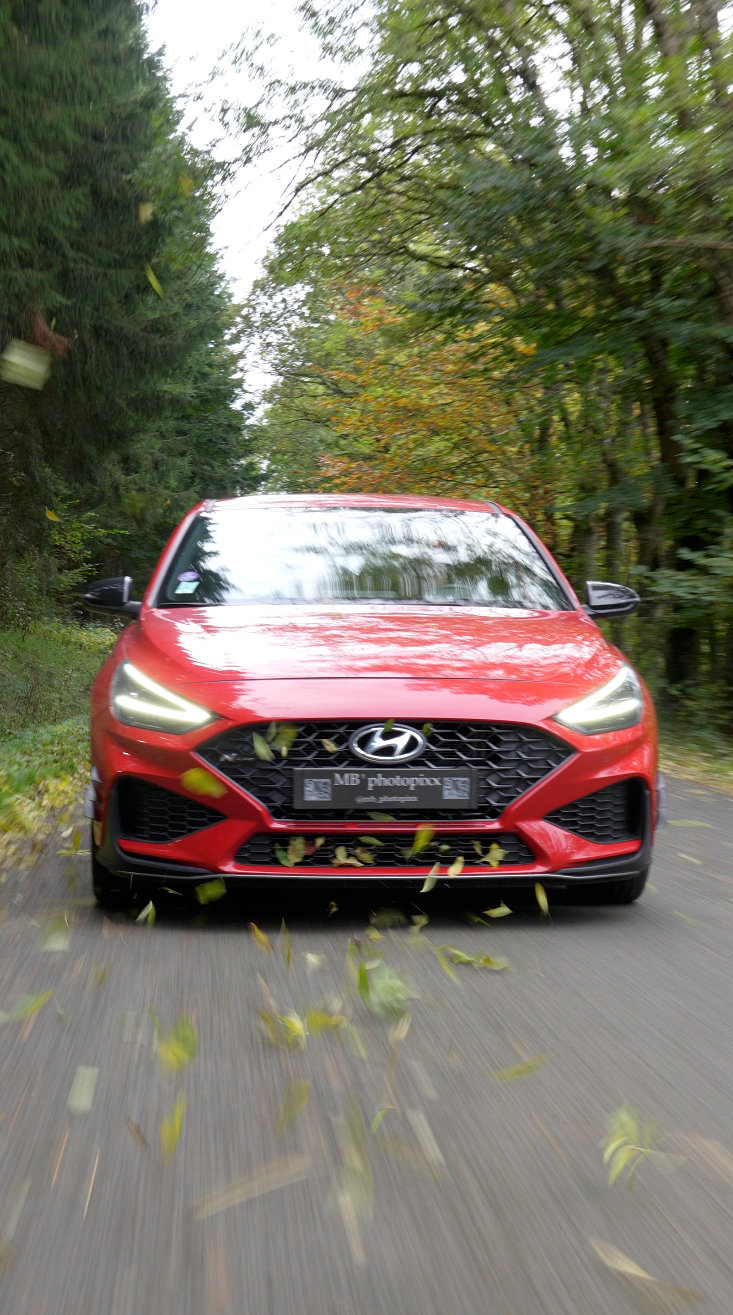 Une voiture rouge en mouvement sur une route entourée d'arbres avec des feuilles qui volent, en forêt.