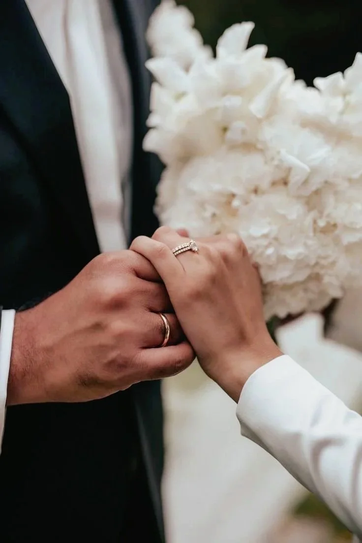 Les mains d'un couple marié tenant leur alliance lors d'une cérémonie de mariage, avec un bouquet de fleurs blanches flou en arrière-plan.