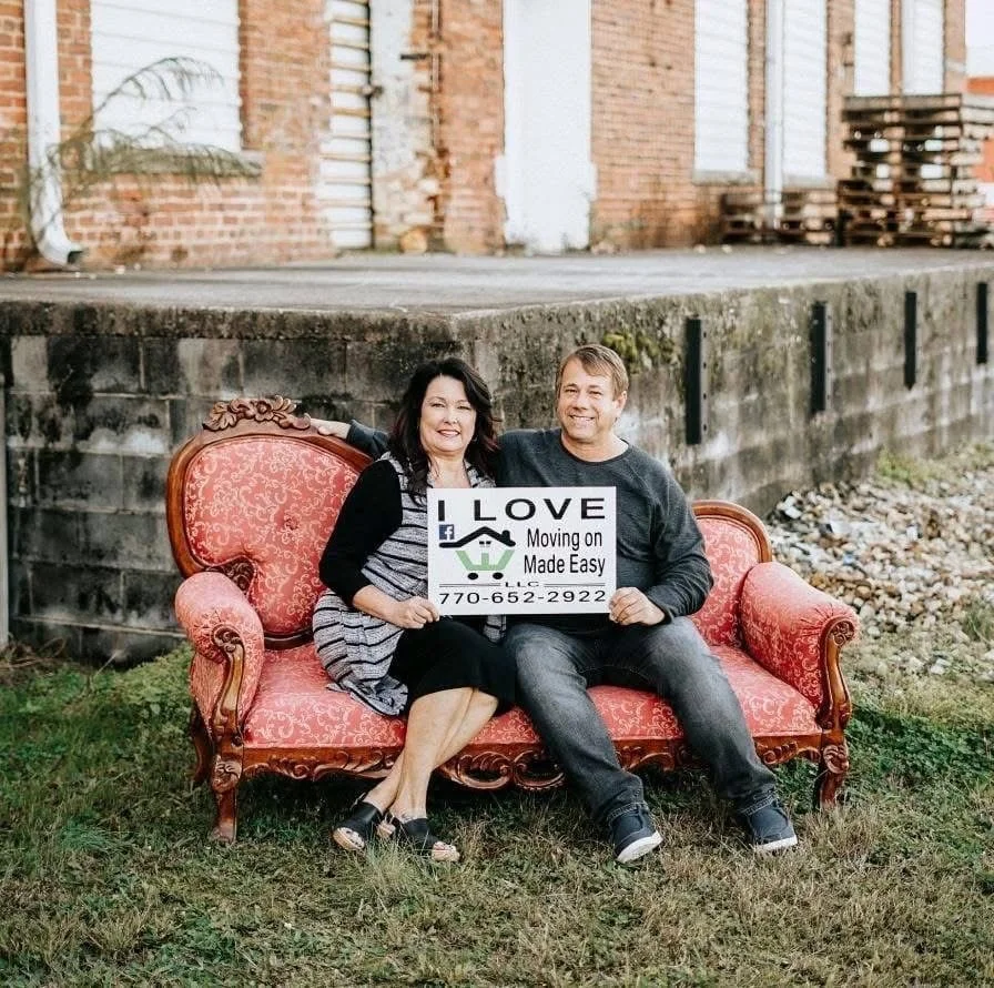 A smiling woman and man sitting on a vintage pink couch outdoors in front of a brick building, holding a sign that says 'LOVE Moving on Made Easy' with a phone number, indicating they are moving or selling a home.