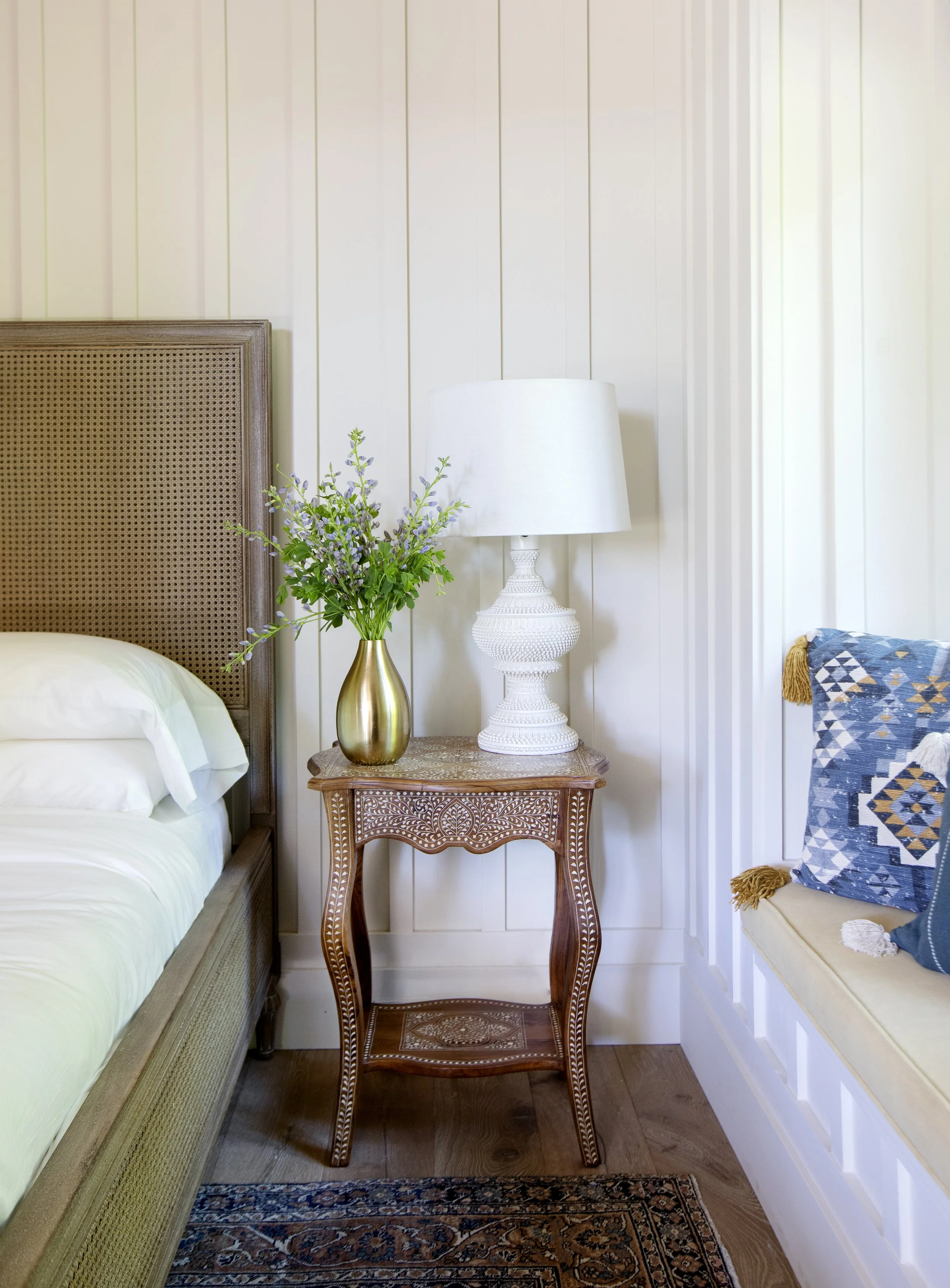 A carved inlay nightstand with a white lamp, a brass vase with purple flowers, a rattan bed with white sheets, an antique persian rug, and a corner of a chair with a blue geometric pillow in a cozy bedroom with shiplap walls.