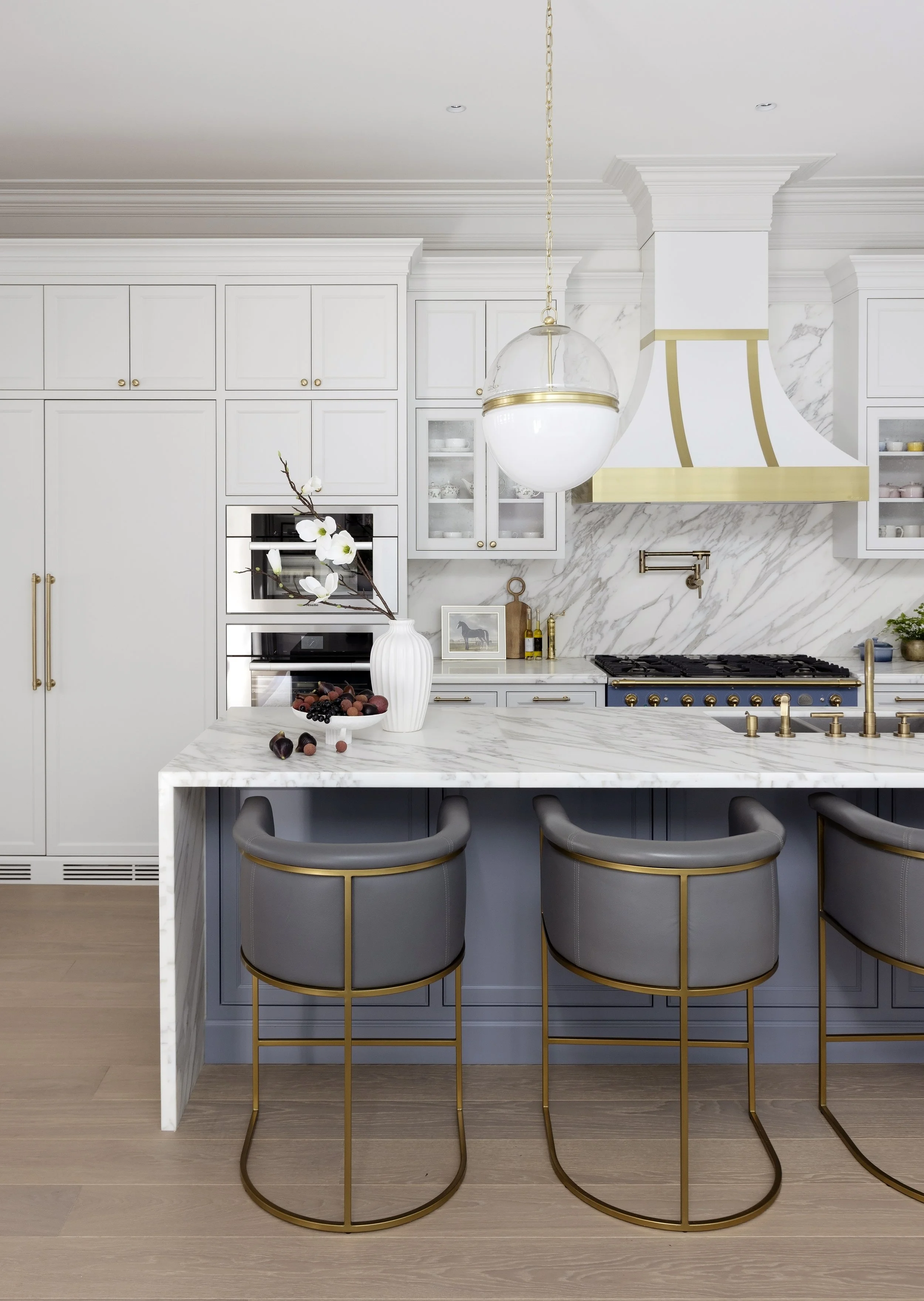 Transitional kitchen with white cabinets, marble backsplash and countertops, brass accents, gray chairs with gold bases, marble island with a vase and fruit, modern gold pendant light.