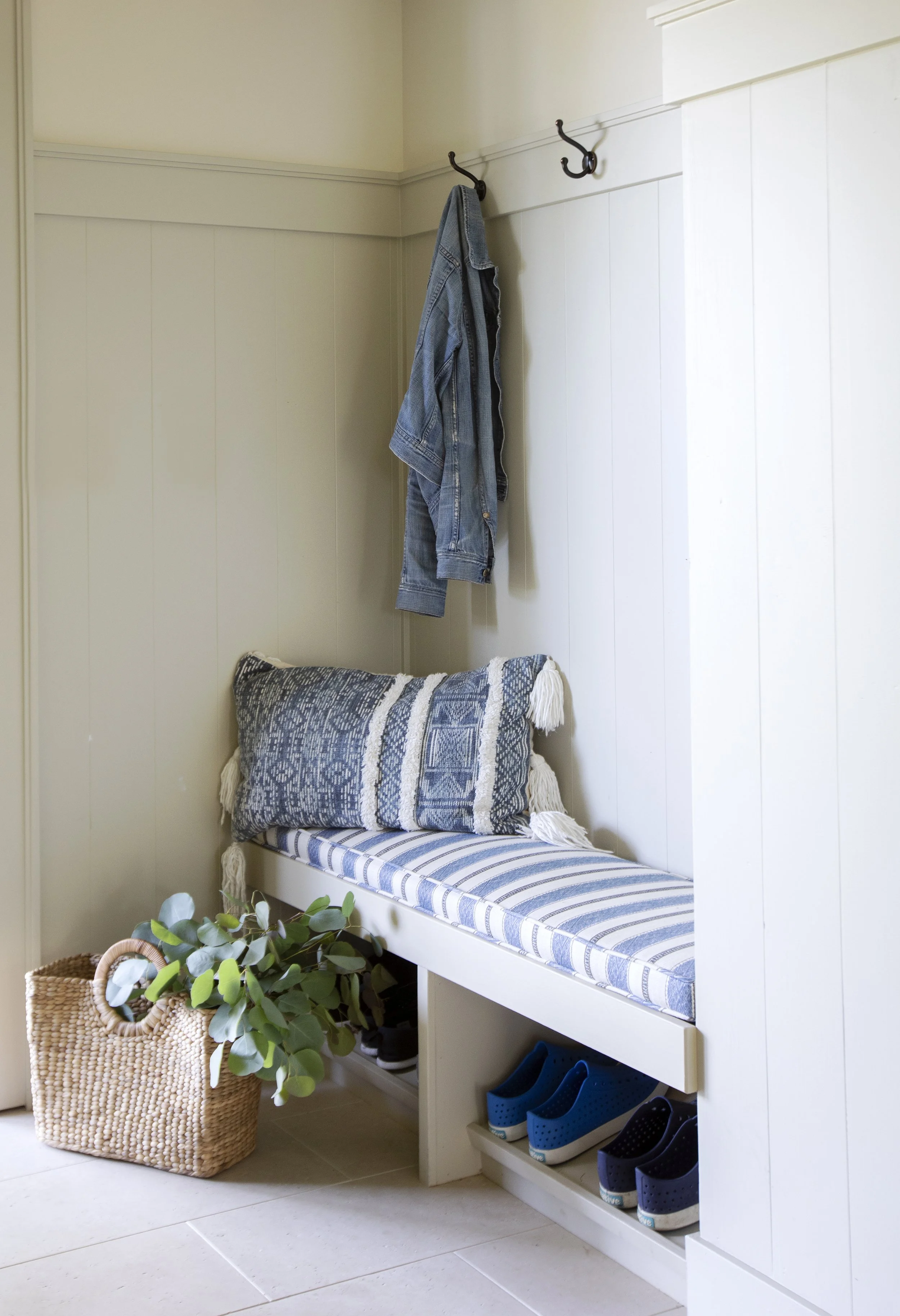 Entryway with a built-in bench, blue and white striped cushion and pillow, hanging denim jacket, basket with green plant, and blue and black shoes arranged in cubbies.