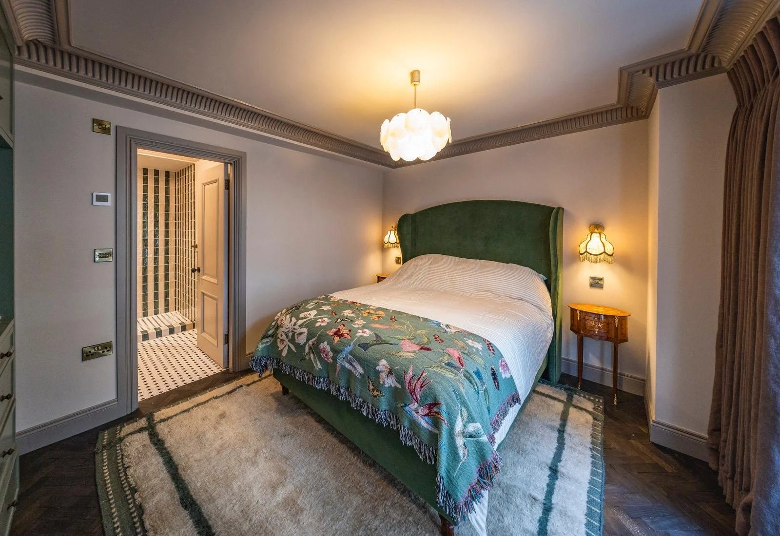 A bedroom with a green upholstered bed, floral blanket, antique french side tables, wall sconces, alabaster chandelier, and an adjacent bathroom with a striped green and white tiled shower.