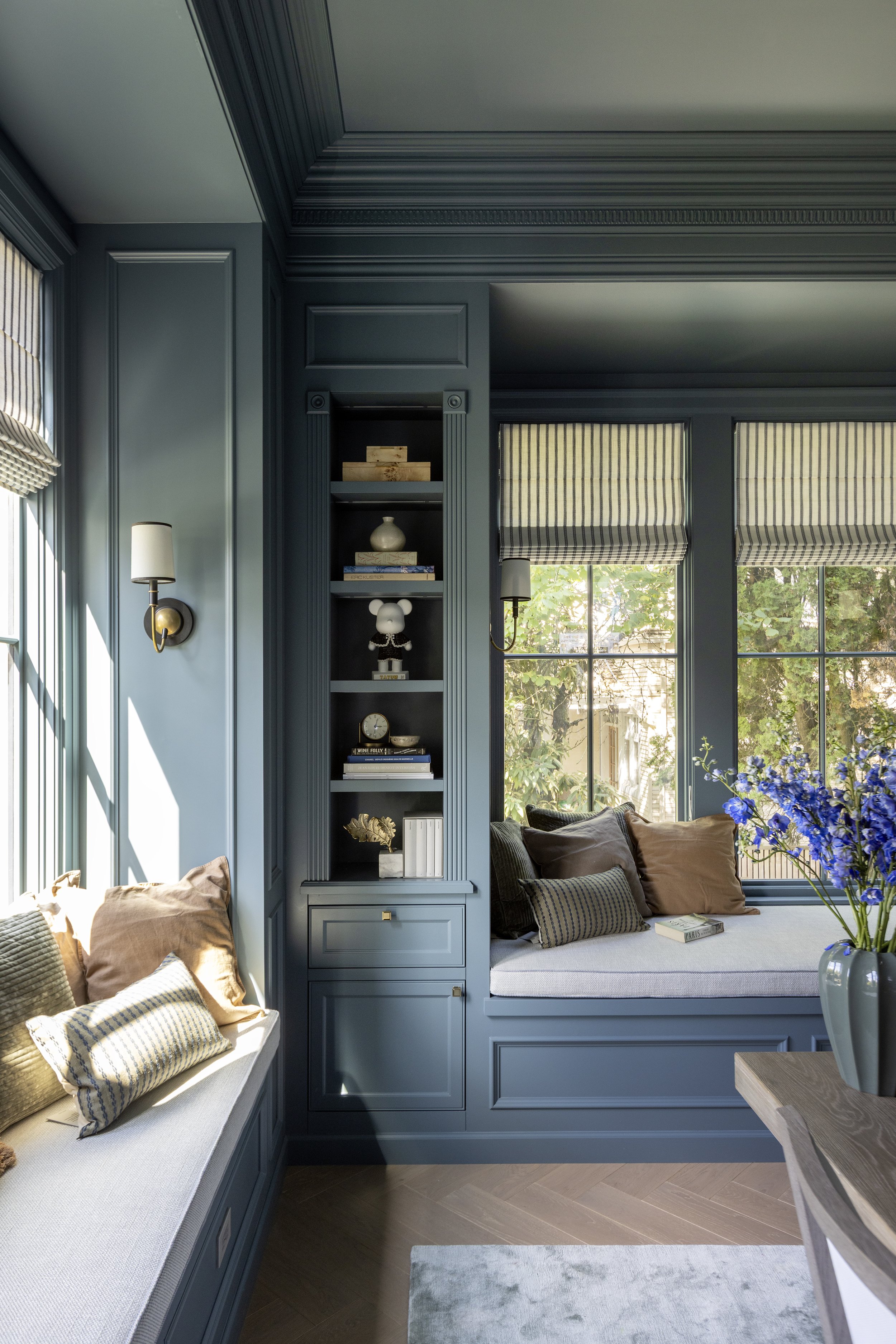 A study with large windows, blue walls, a cosy chenille window seat with beige and striped cushions, books and decor, built in bookcase with decorative items, and a smoked grey wood desk with a blue rug.