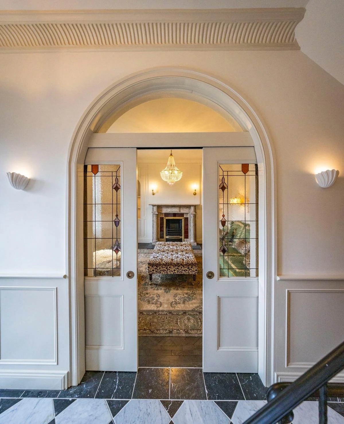 Interior view of a living room through arched double doors with stained glass panels, featuring a fireplace, chandelier, and leopard print ottoman.