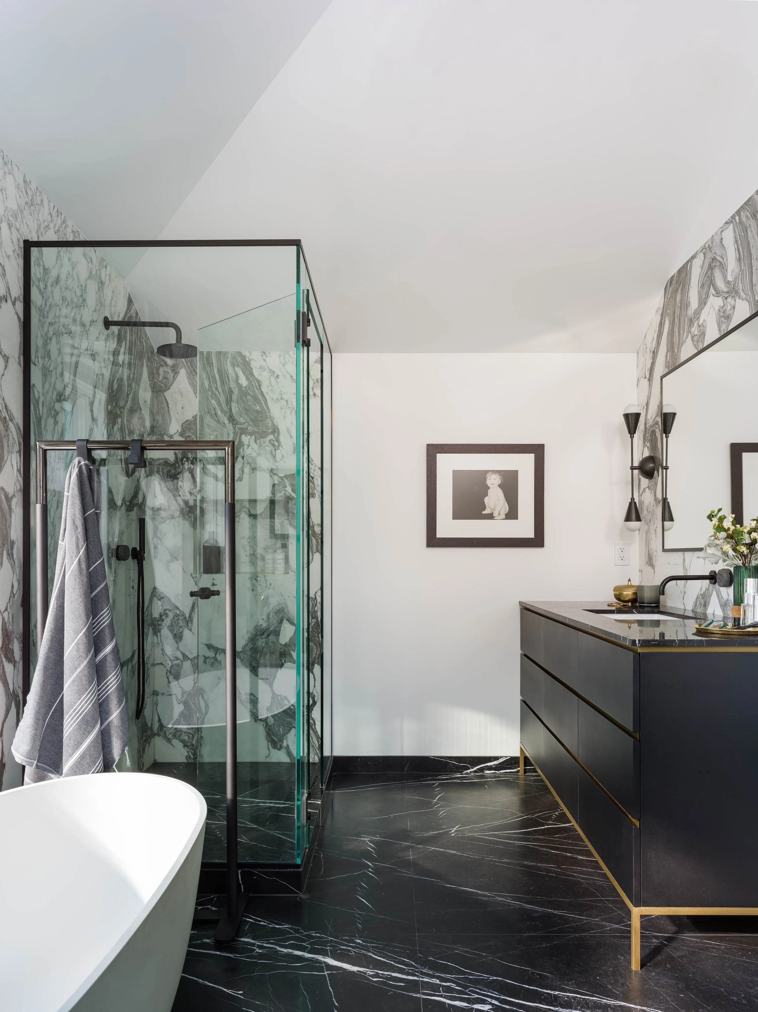 Modern bathroom with a black marble floor, a white bathtub, a glass-enclosed shower with marble walls, black fixtures, and a black vanity with brass accents. Framed artwork and black wall sconces are visible.