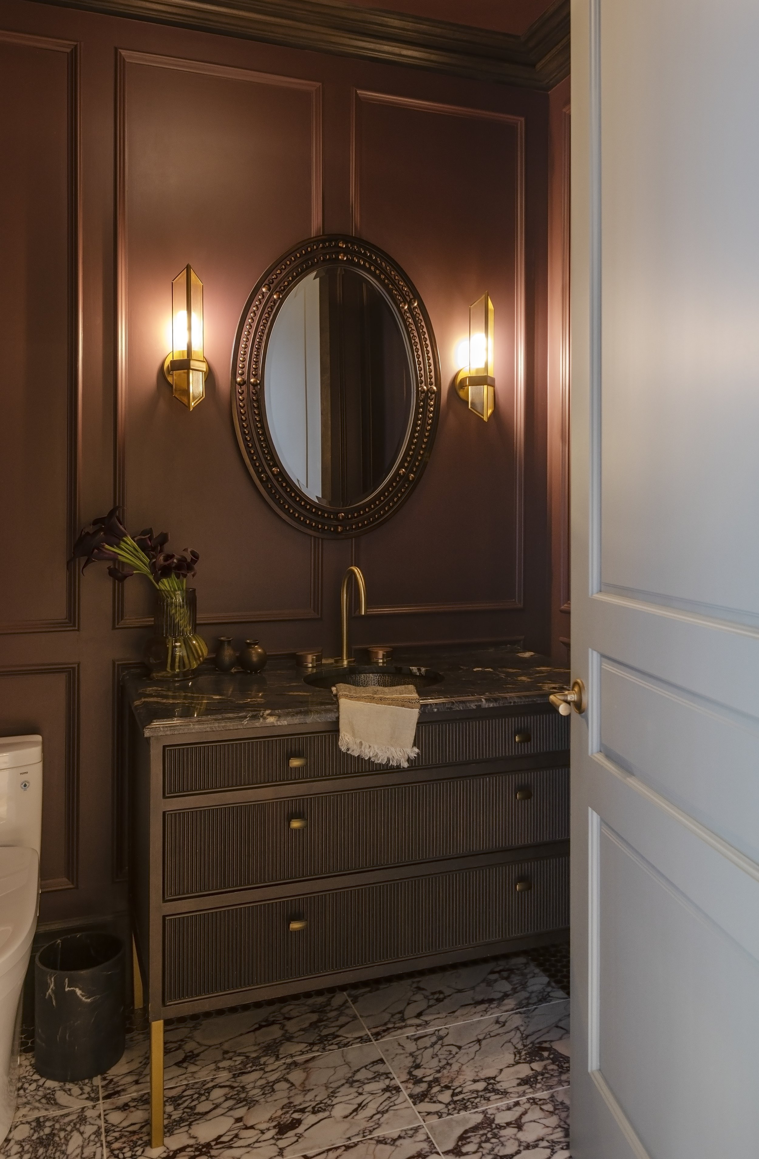 A bathroom with merlot paneled walls, an oval mirror with a beaded frame, reeded glass wall sconces, a marble countertop with a brass faucet, a vase with dark lilies, and burgundry marble floor. A green door is open, revealing the room.