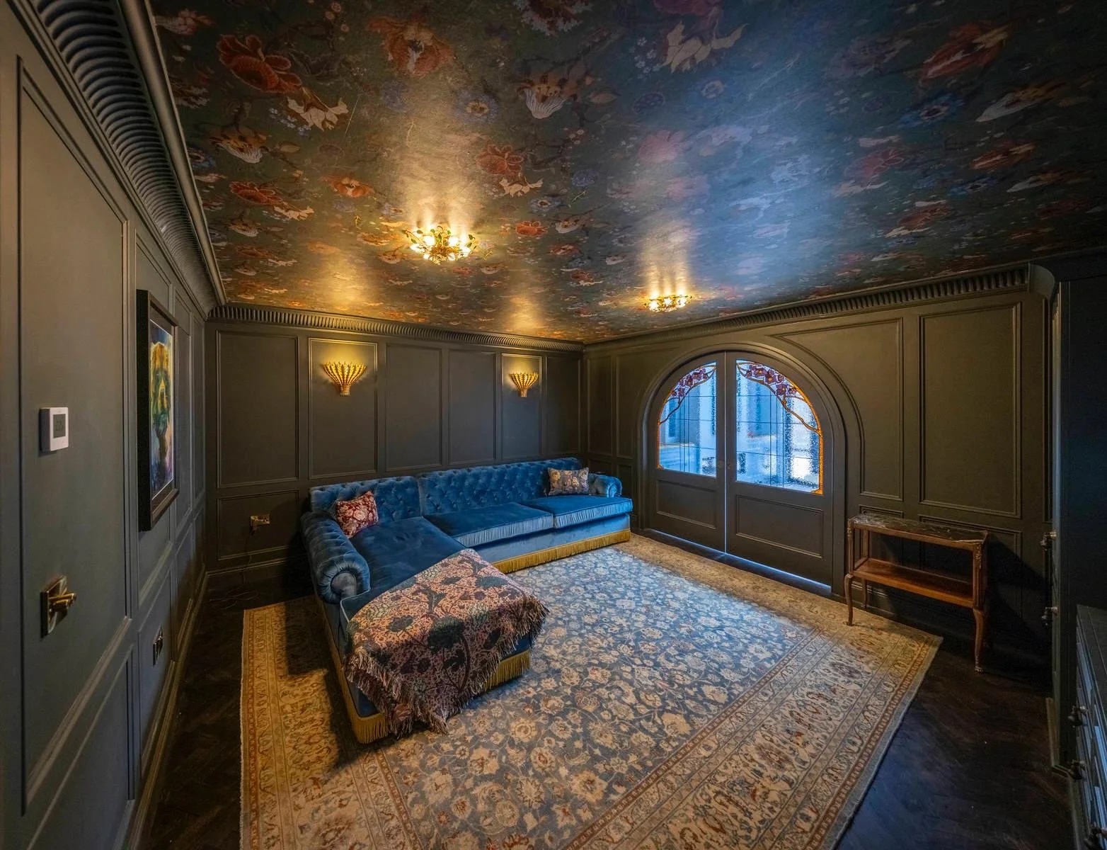 A luxury lounge with dark green paneled walls, a floral patterned ceiling, and vintage wall sconces, featuring a blue velvet tufted sofa, an antique persian rug, stained glass doors, and a small wooden table near the window.