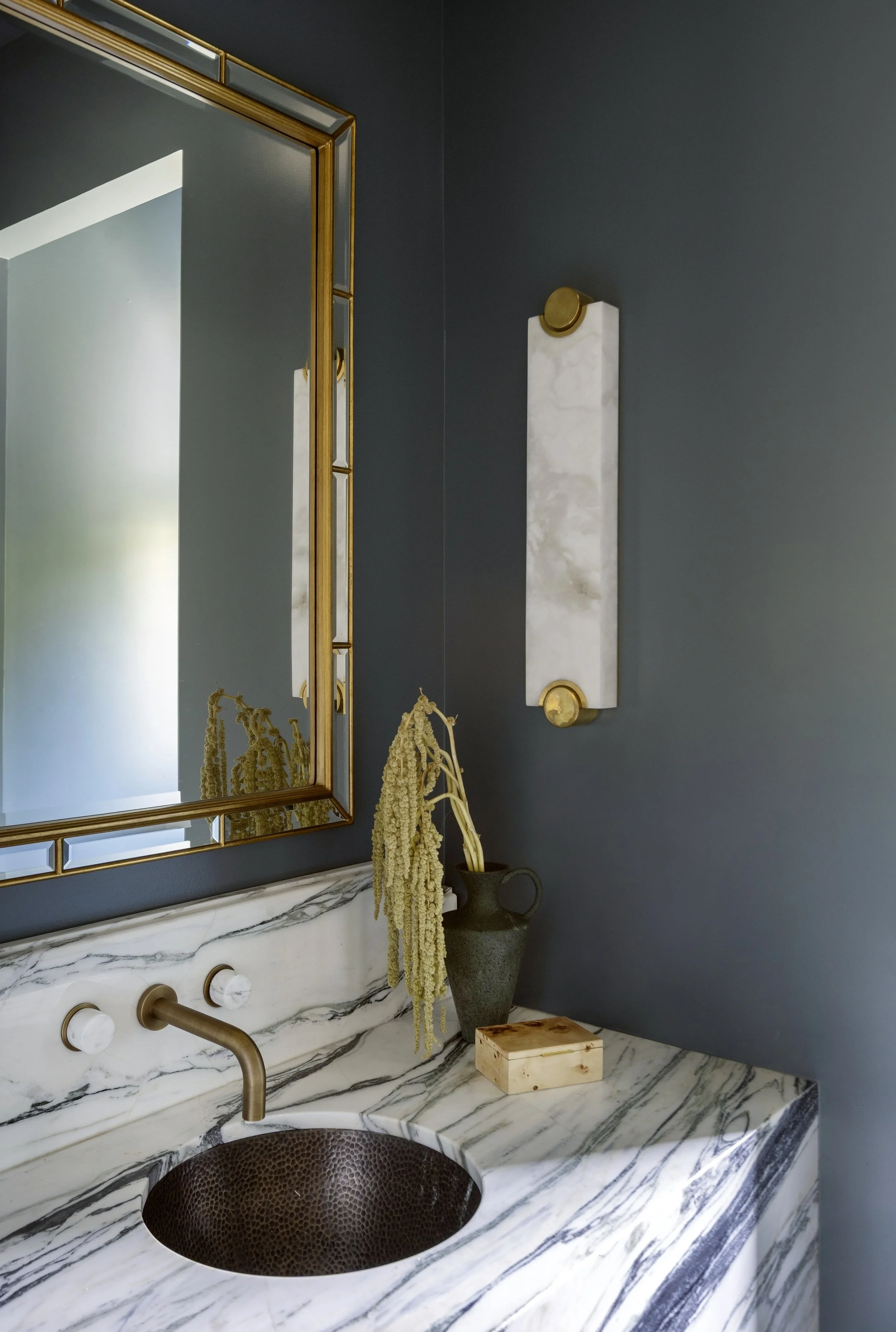 A bathroom vanity with a marble countertop, mounted bronze faucet, and textured bronze sink. A large mirror with a gold frame is above, with dark blue walls. Alabaster wall sconces, a vase with draped flowers, and a burl wood box on the counter.