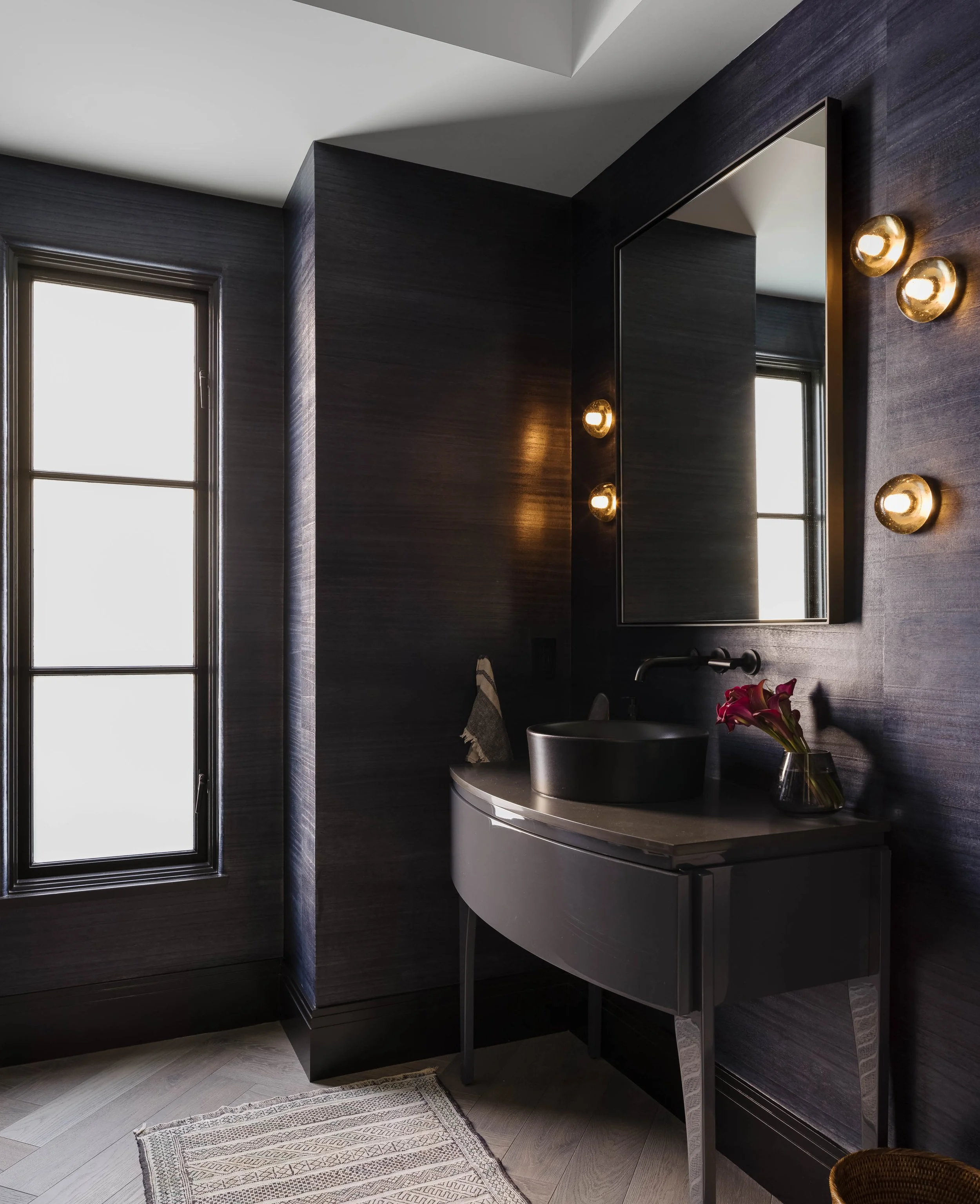 Modern bathroom with a lacquered vanity, and smoked glass wall sconces . A vessel sink and a vase of burgundy lilies on the countertop. Natural light streams in through a tall window, onto navy wood wallcovering.