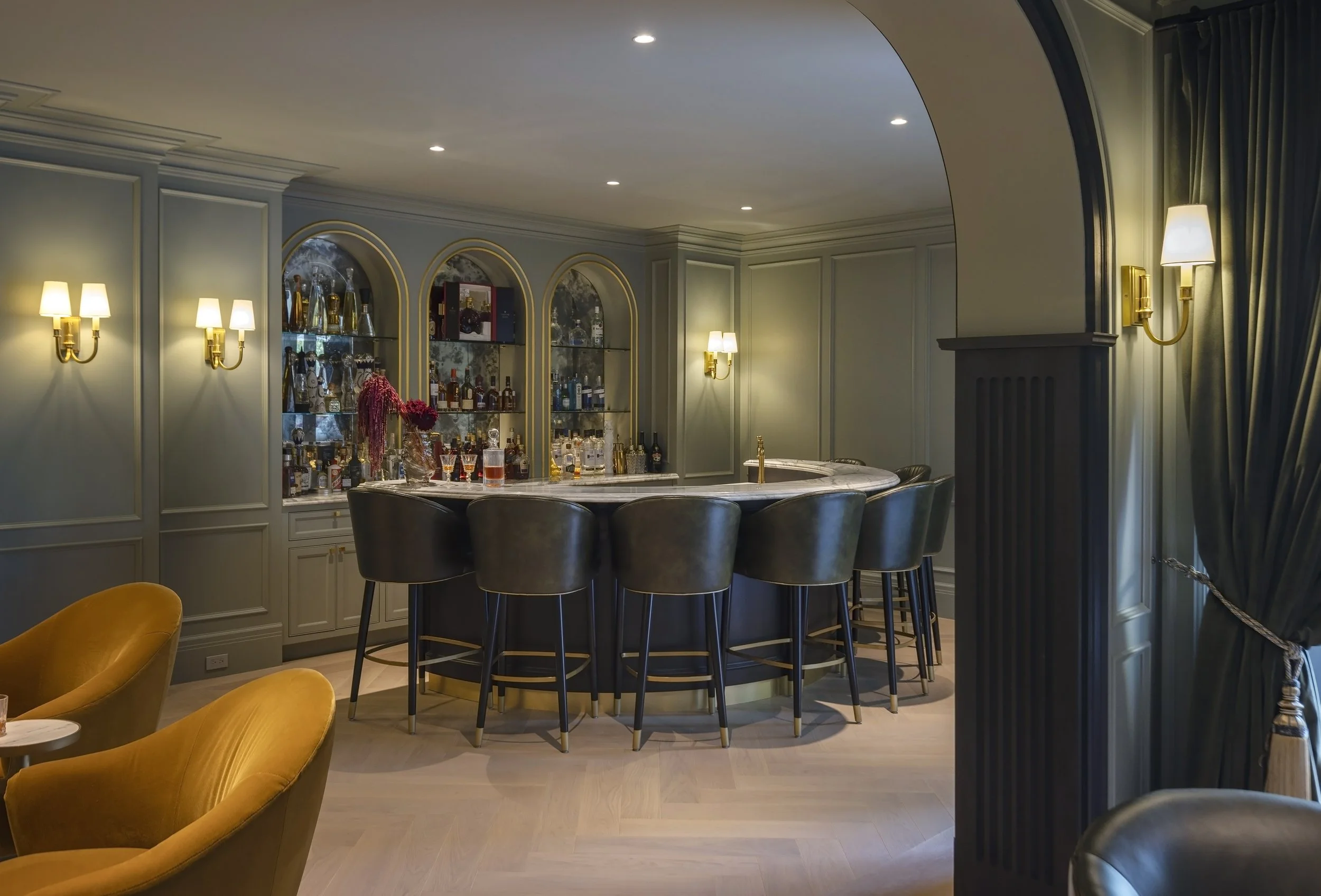 An elegant home bar with a marble countertop, surrounded by dark barstools, with a well-stocked liquor shelf behind it. The room features soft lighting and soft green walls with brass accents.