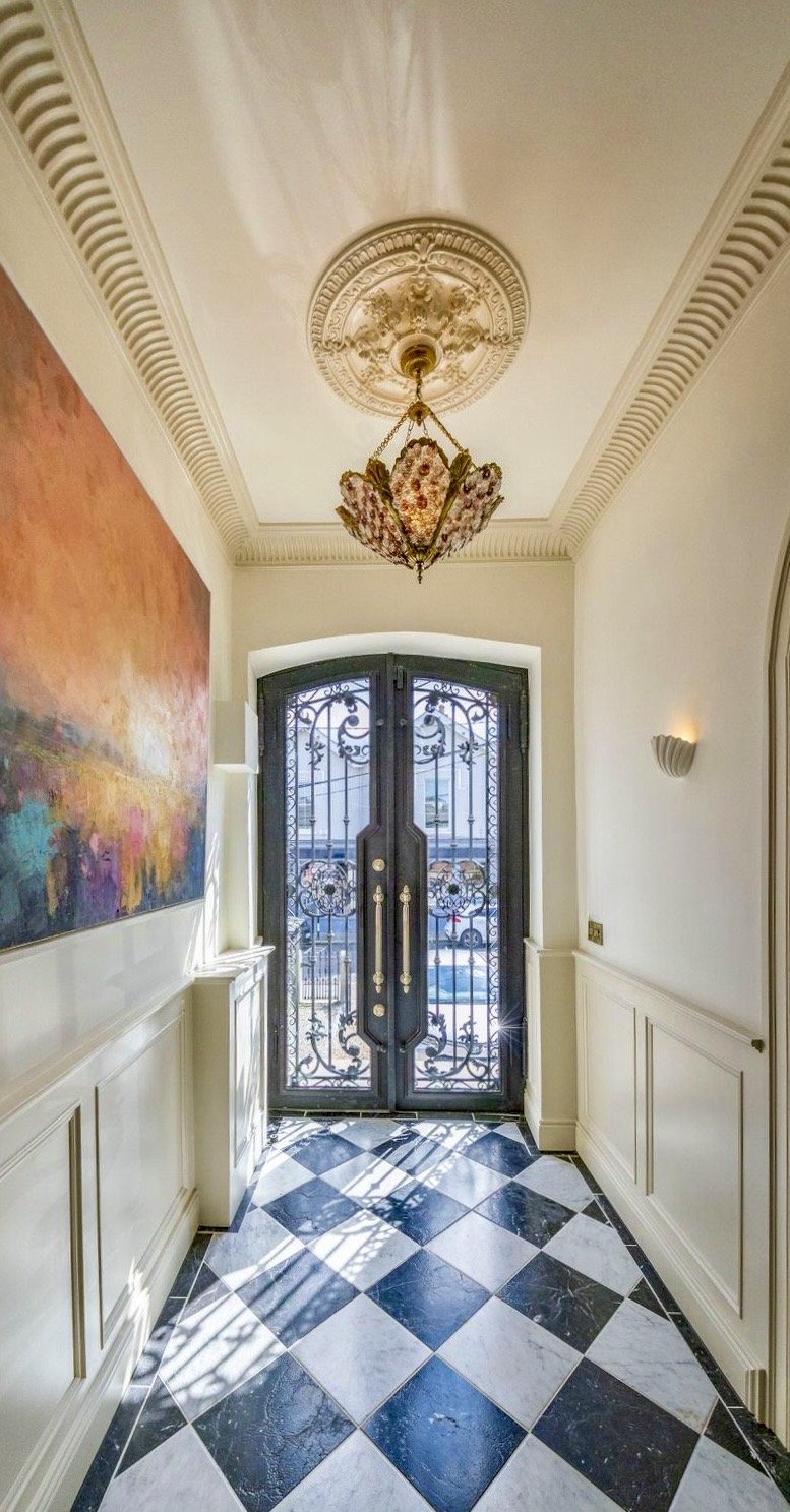 Elegant entryway with black ornate double doors, a floral chandelier, colorful abstract wall art, and a checkered black and white marble floor.