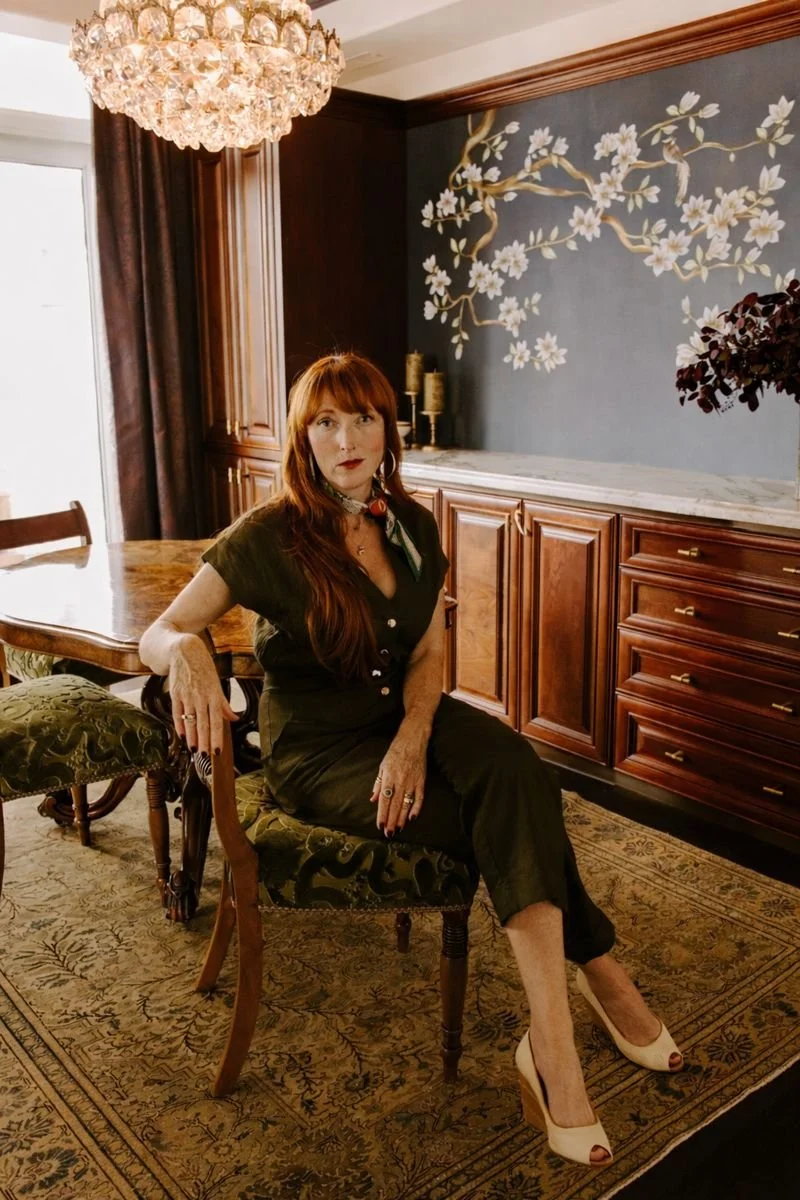 A woman with red hair sitting on an ornate chair in a dining room with dark wood furniture, a chandelier, and floral wall art.