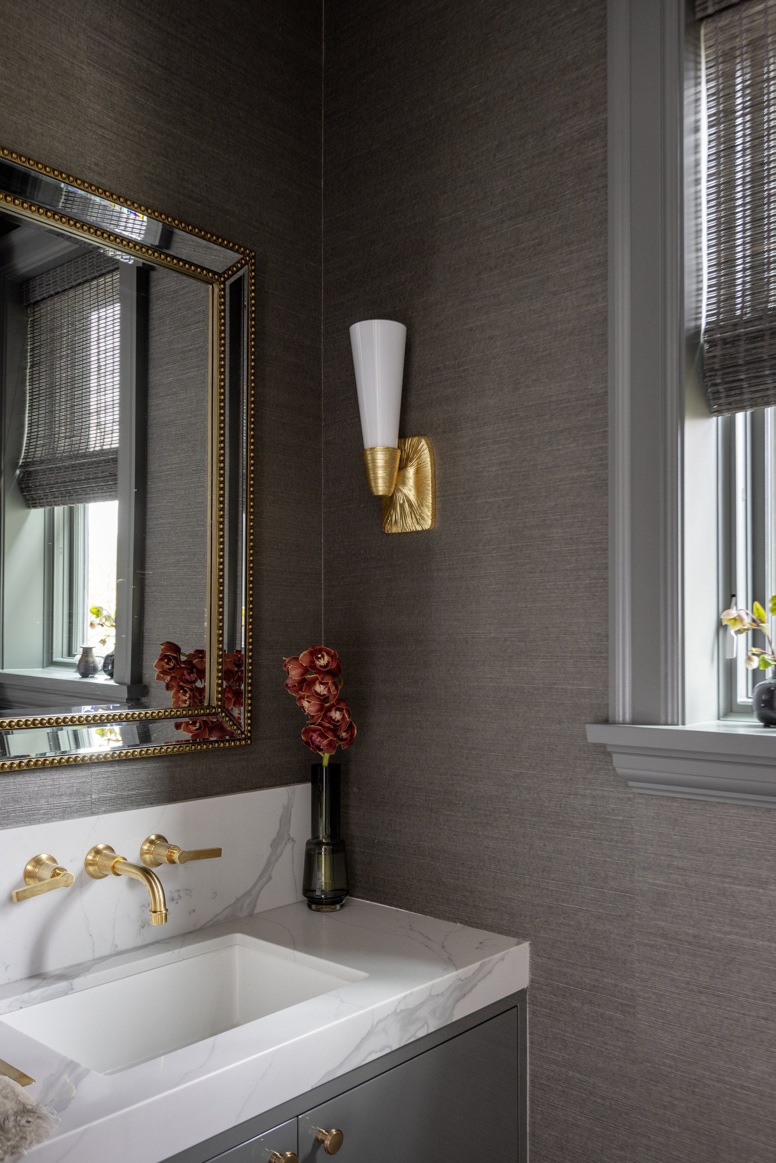 A bathroom vanity with a white marble sink and gold fixtures, a large mirror with a gold beaded frame, a wall sconce with a gold base and white shade, a tall black vase with red flowers, and windows with blinds.