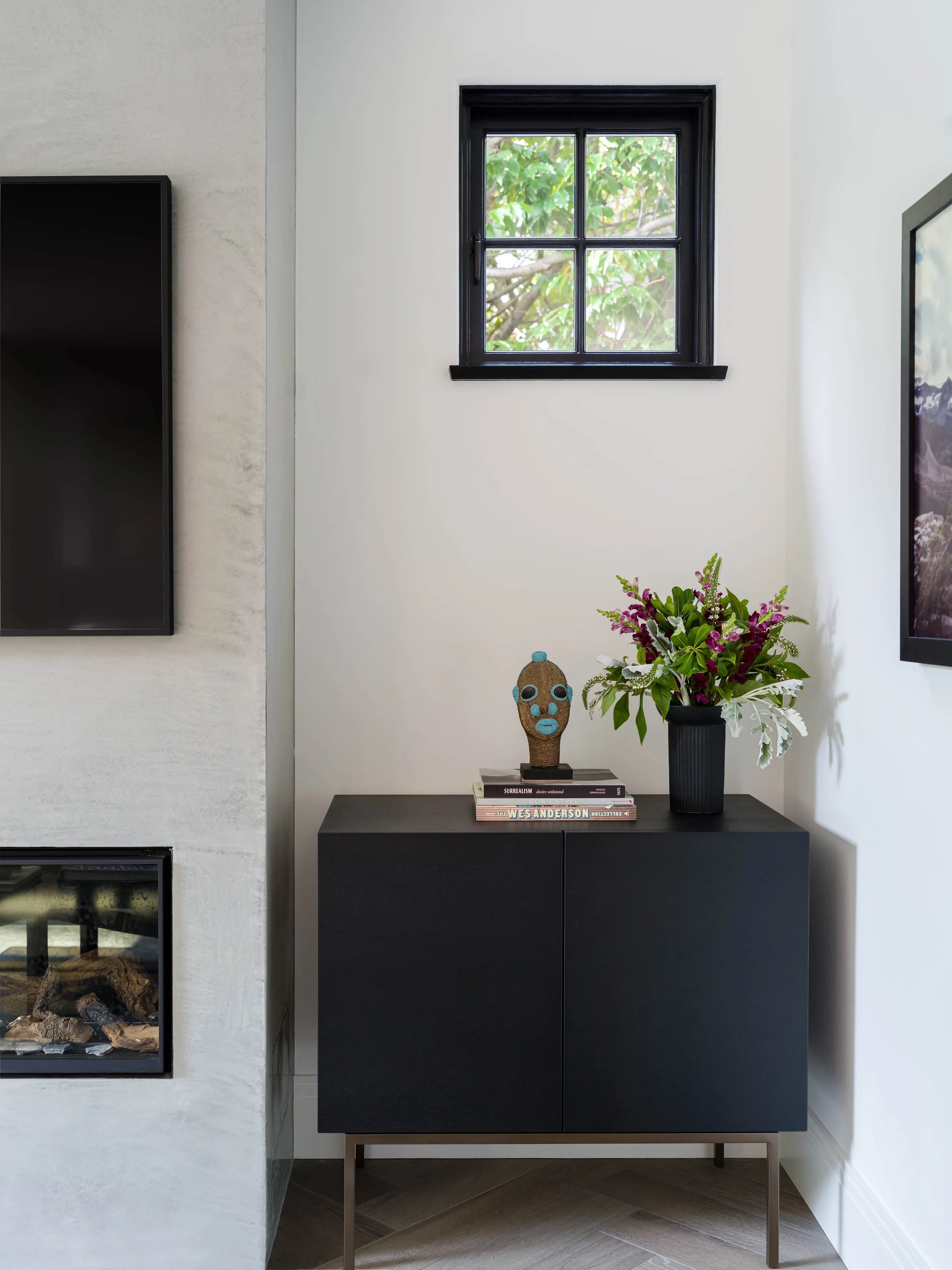 Interior of a modern room with concrete fireplace mantel, a black stained wood cabinet with brass legs, a small window showing green foliage, a colorful flower arrangement beside an artisan beaded sculpture, and decorative books.
