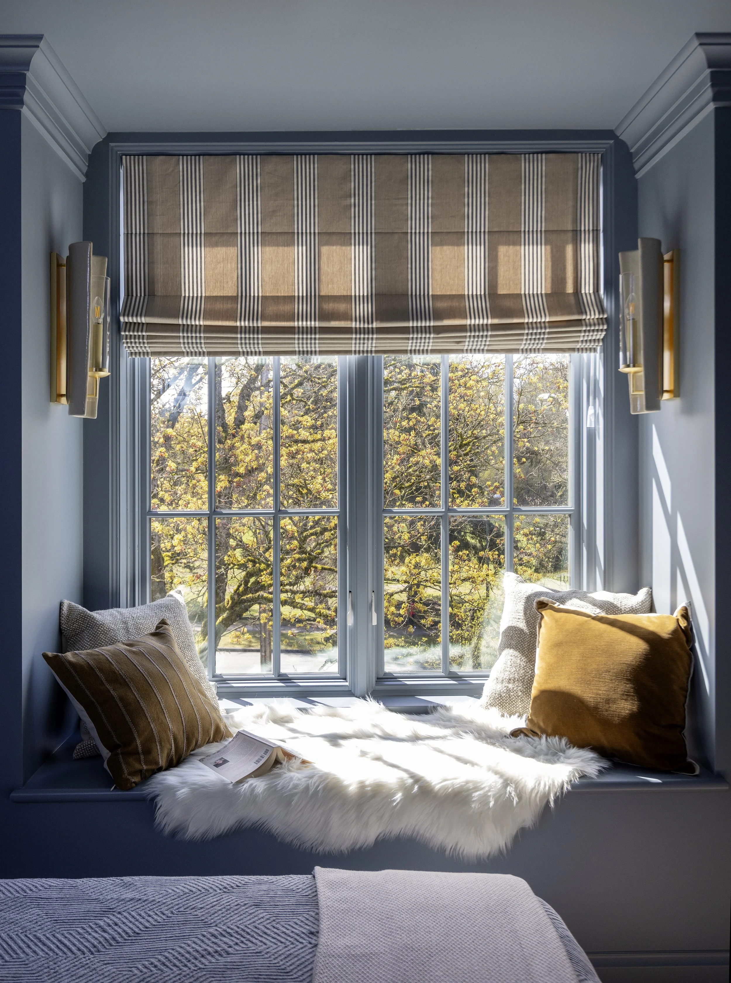 Sunlit cozy window nook with striped Roman shades, glazed porcelain wall sconces, beige and ochre pillows, a furry white blanket, and a view of trees outside.