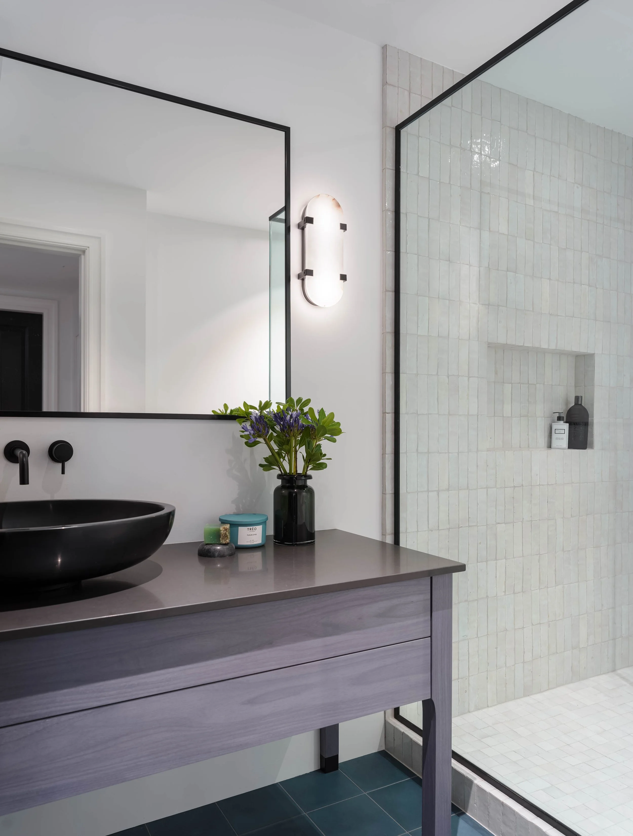 Modern bathroom with a blue tiled floor, a black vessel sink on a stained wood blue vanity, a large mirror, alabaster wall sconce, and a walk-in shower with white zellige tiles separated by a glass partition.